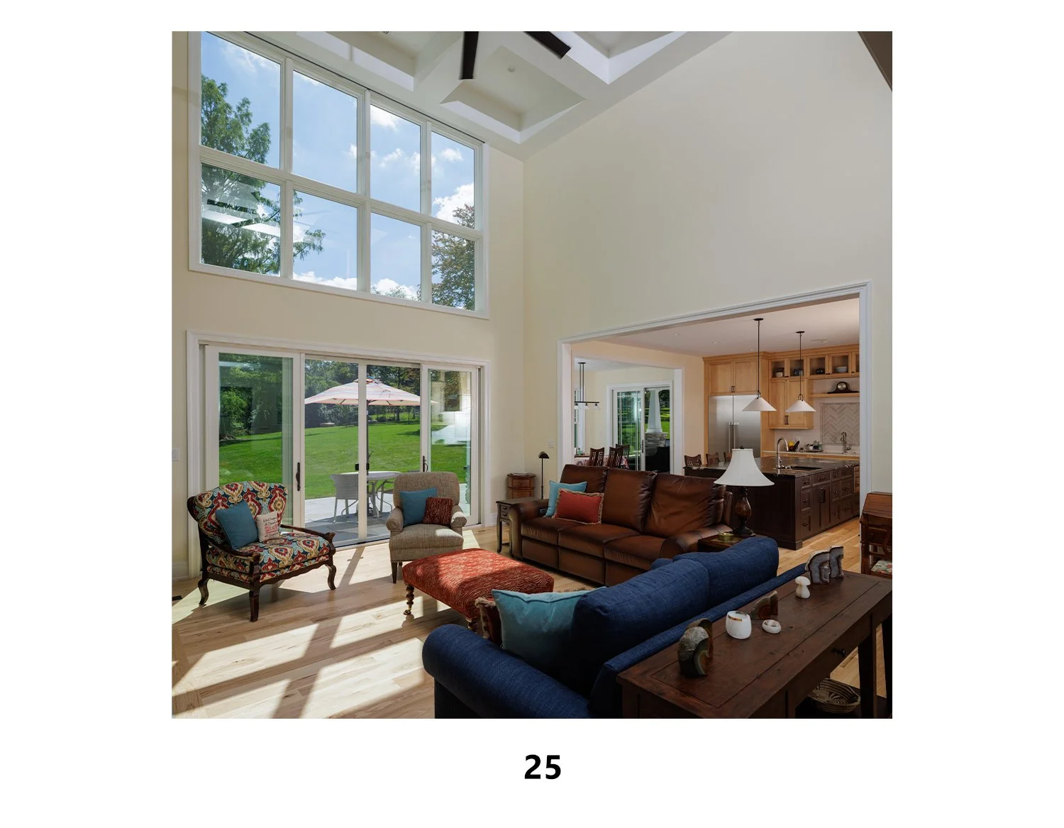 Living room with tall windows showcasing a grassy backyard with outdoor furniture, a brown leather sofa, patterned armchairs, a blue allover couch, a sliding glass door, and a view into the kitchen with wooden cabinets and a stainless steel refrigera