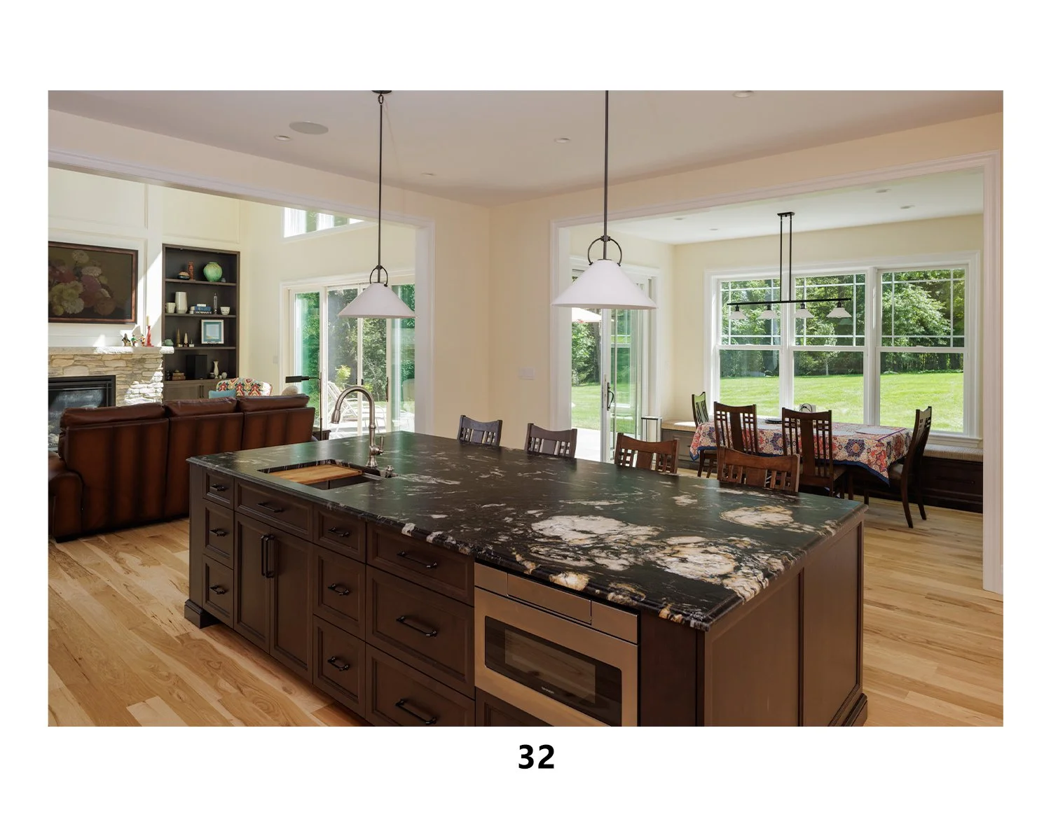 Open-concept kitchen and dining area with large windows overlooking a green backyard, a marble kitchen island with a sink, pendant lighting, a dining table with a colorful tablecloth, and a cozy living room with a fireplace and built-in shelves.