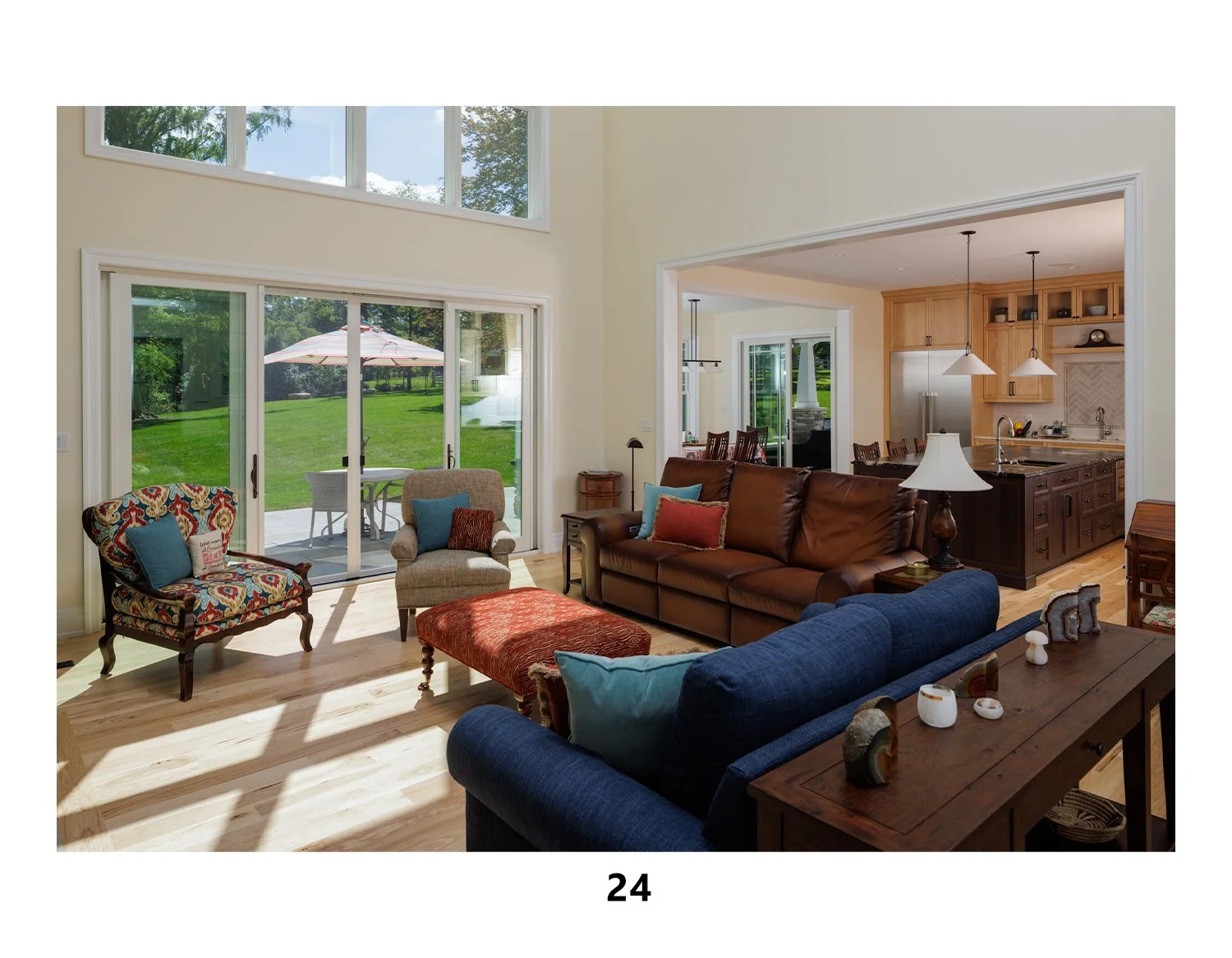 Living room with large sliding glass doors leading to a green backyard, featuring various chairs, a brown leather sofa, a blue sofa, and a wooden table, with a view into the kitchen area.