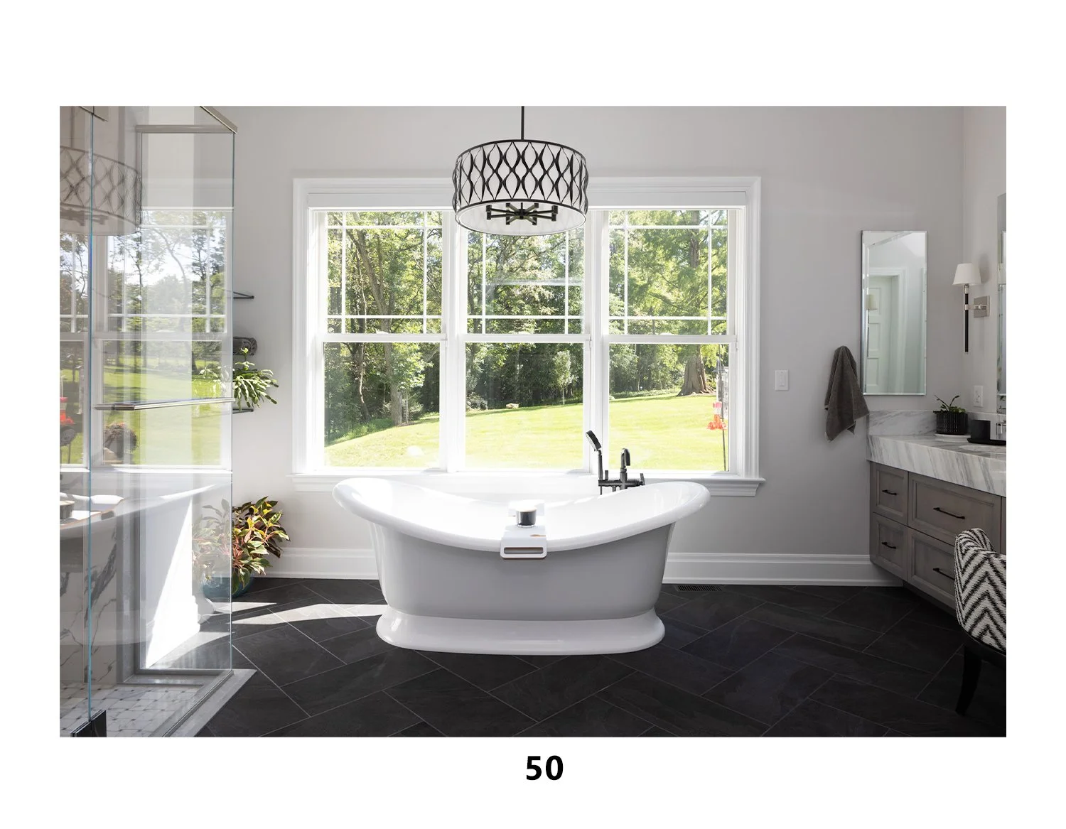 Modern bathroom with a white bathtub, large window showing a green outdoor landscape, gray vanity with a mirror, and black flooring.