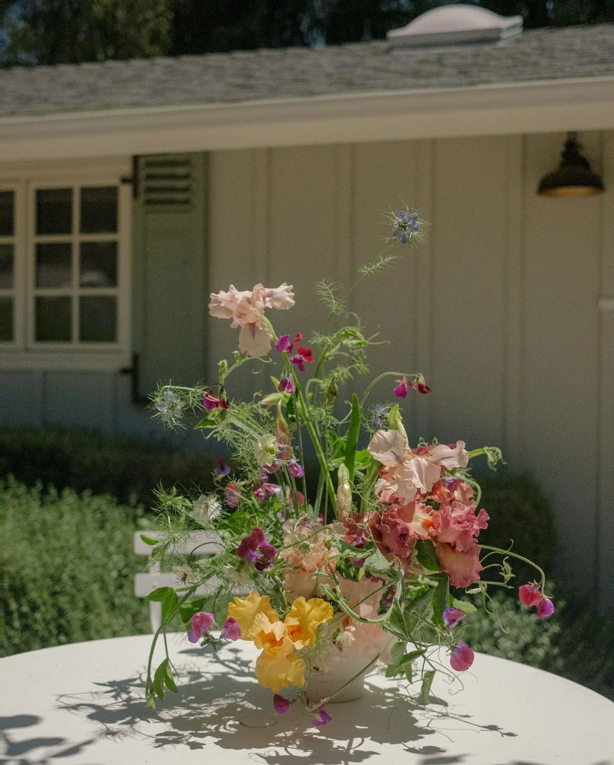 A colorful flower arrangement in a white vase on a white table outside, with a house and greenery in the background.