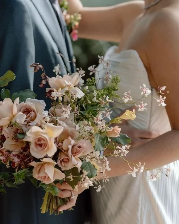 Close-up of a bride and groom holding a bouquet of pink and cream flowers at a wedding, with the bride wearing a white dress and the groom in a dark suit.