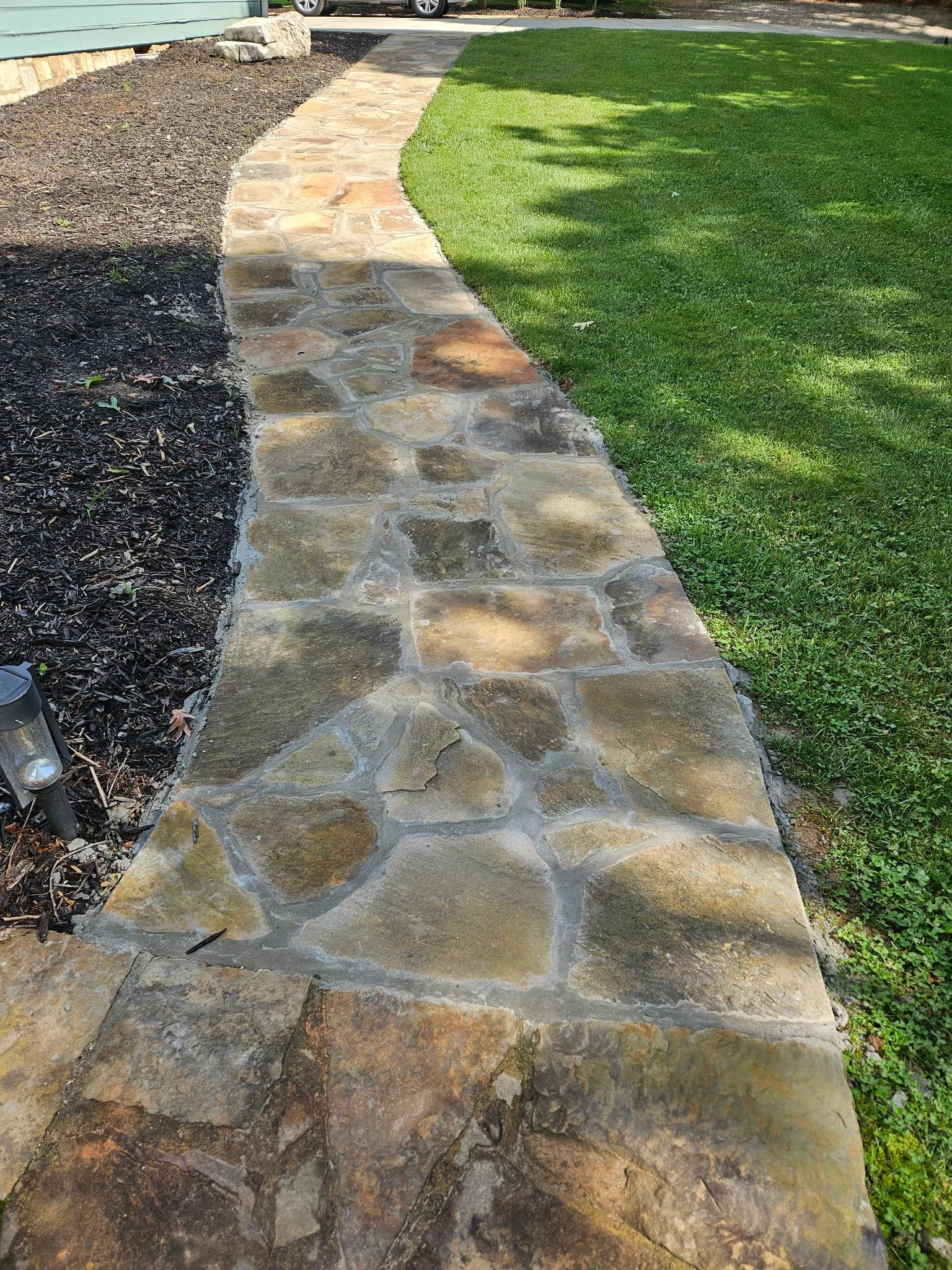 A curved stone pathway with large, irregularly shaped stones leading through a landscaped yard with green grass and a bed of dark mulch.