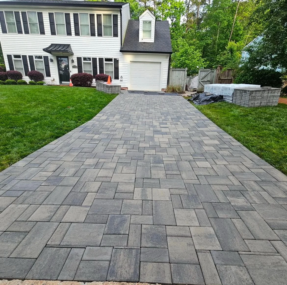 Newly paved stone driveway leading to a white two-story house with black shutters and a garage, surrounded by green grass and trees.