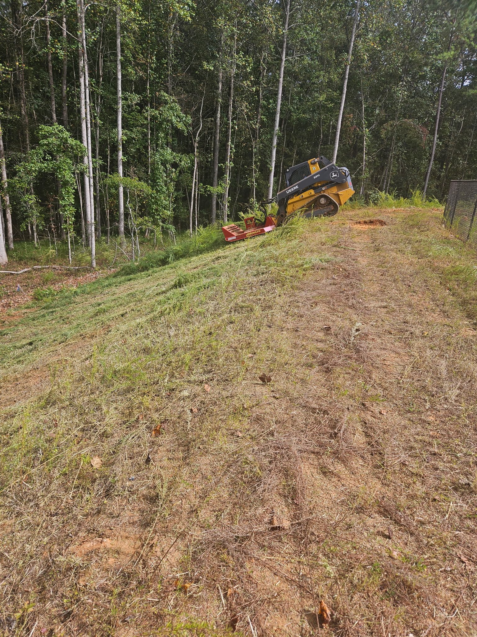 A small yellow and black John Deere compact excavator parked on a sloped grassy area near a wooded forest.