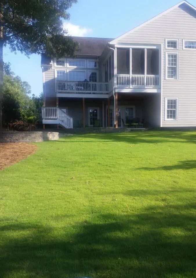 A two-story house with beige siding and white railings, surrounded by a well-maintained green lawn and trees, under a partly cloudy sky.