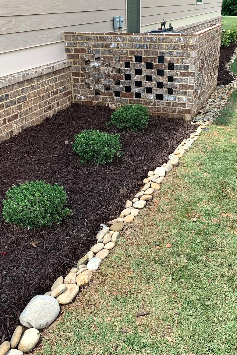 A landscaped garden with three trimmed bushes, dark mulch, a border made of smooth light-colored rocks, and a brick wall with some open brickwork on the right side.