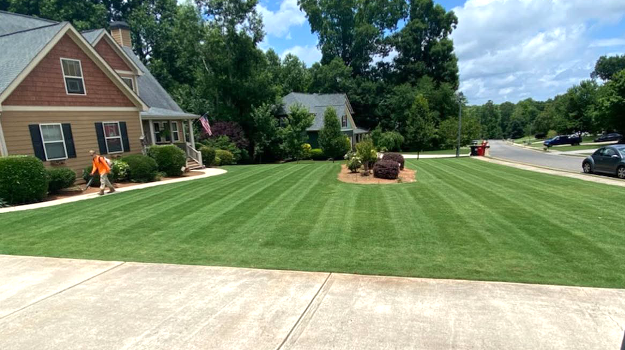 Well-maintained grassy lawn with striped pattern, surrounded by landscaping with bushes and trees, near residential houses with parked cars on the street, under partly cloudy sky.