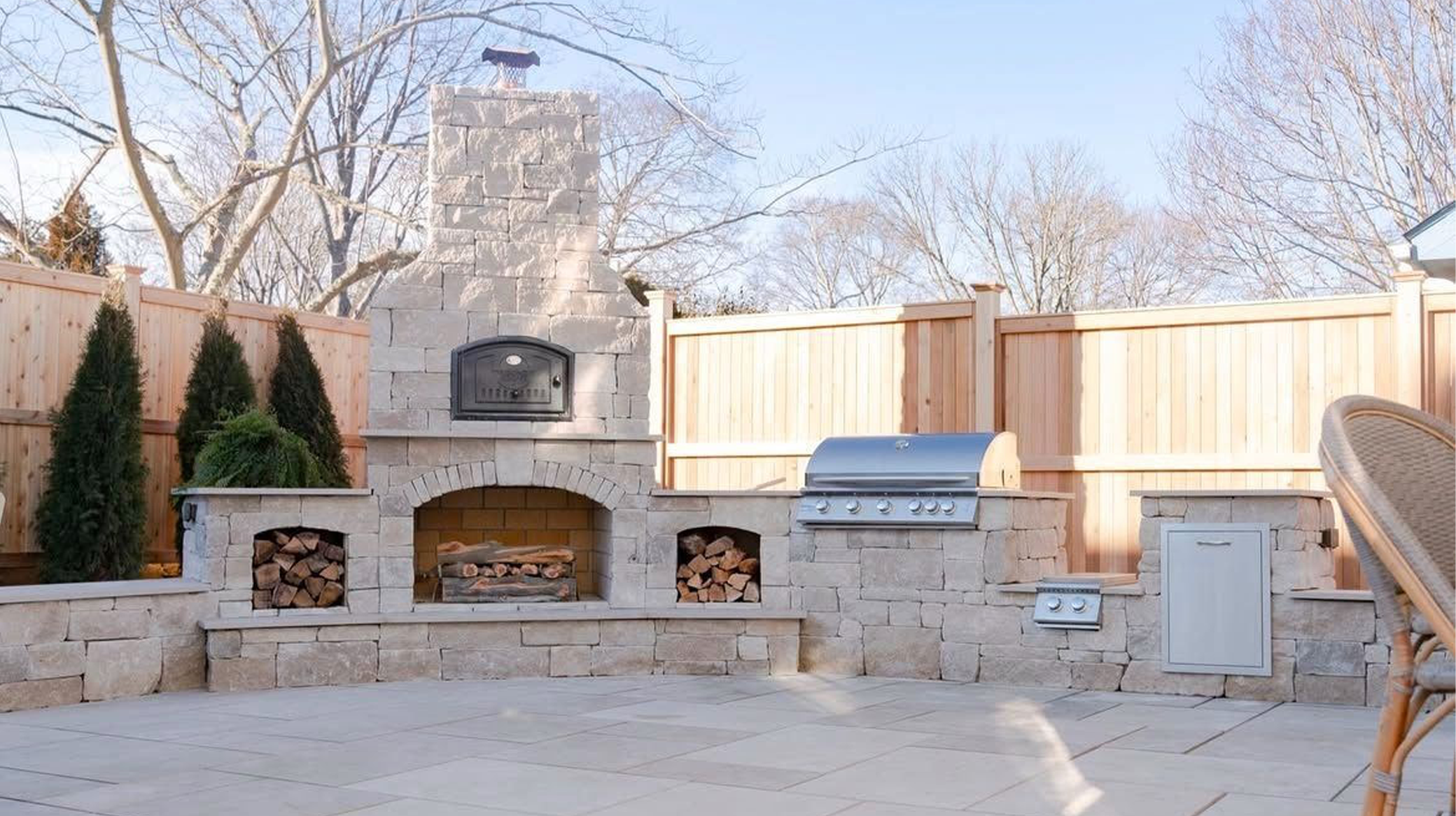 Outdoor backyard kitchen with a stone fireplace, wood storage, a stainless steel grill, and a wooden fence.
