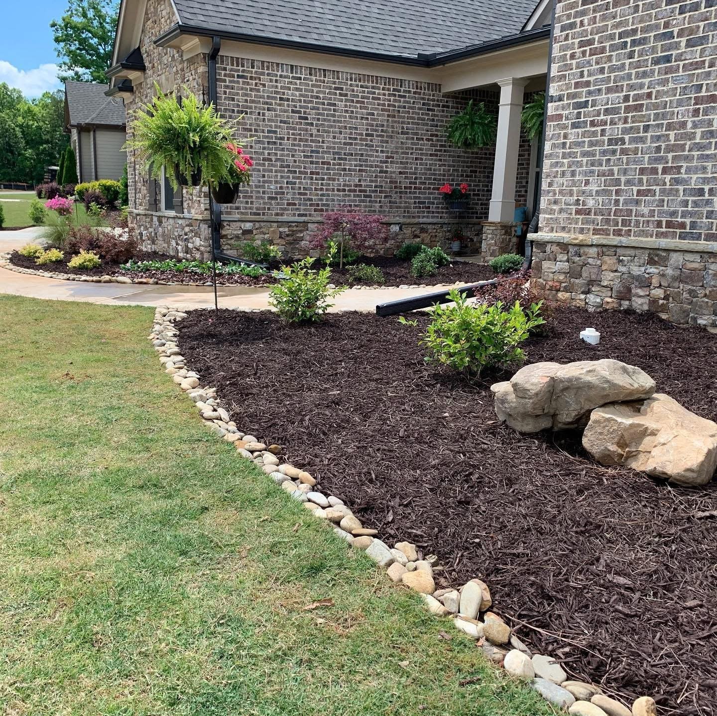 Landscaped front yard with a brick house, mulched flower beds with small shrubs and rocks, and a curved walkway with a hanging plant.