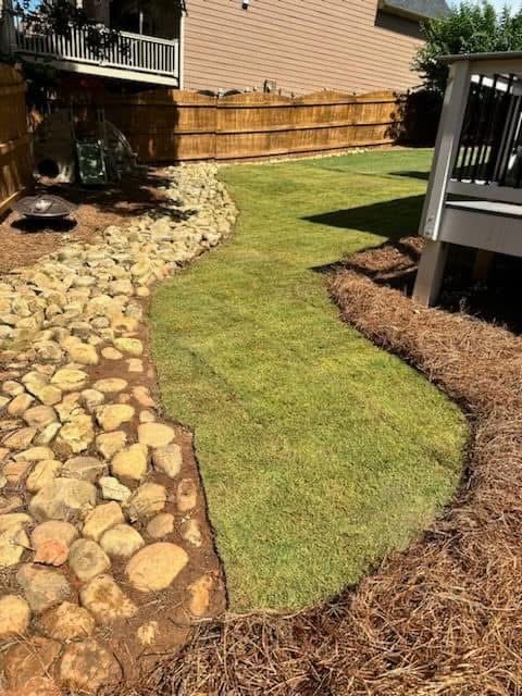 A backyard with a manicured grass lawn, a rock border on the left, and a mulch border on the right. There is a wooden fence in the background and part of a deck on the right side.