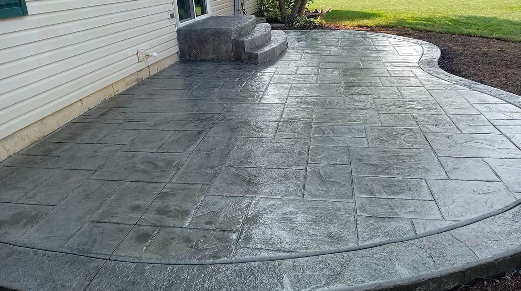 A freshly installed concrete patio with stamped and stained pattern, adjacent to a house with siding and concrete stairs leading to a door, with a lawn in the background.