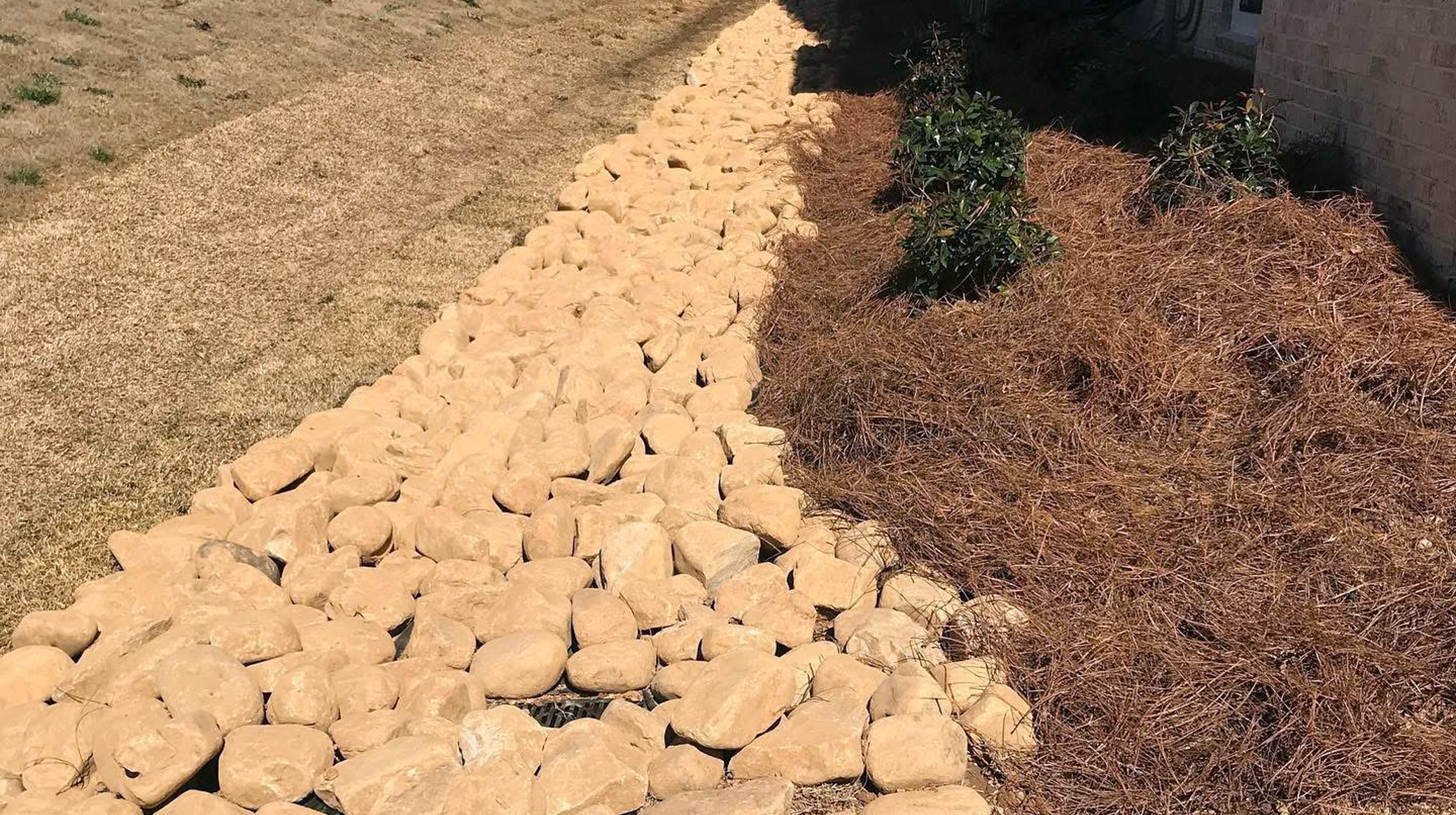 Rock mulch border alongside a garden bed with bushes, adjacent to a brick house wall and a lawn.