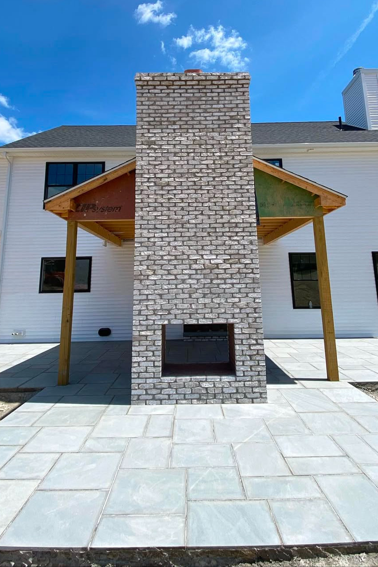 Photo of a house exterior featuring a brick chimney, a covered patio with wooden supports, and a white siding house with black window frames under a blue sky with clouds.