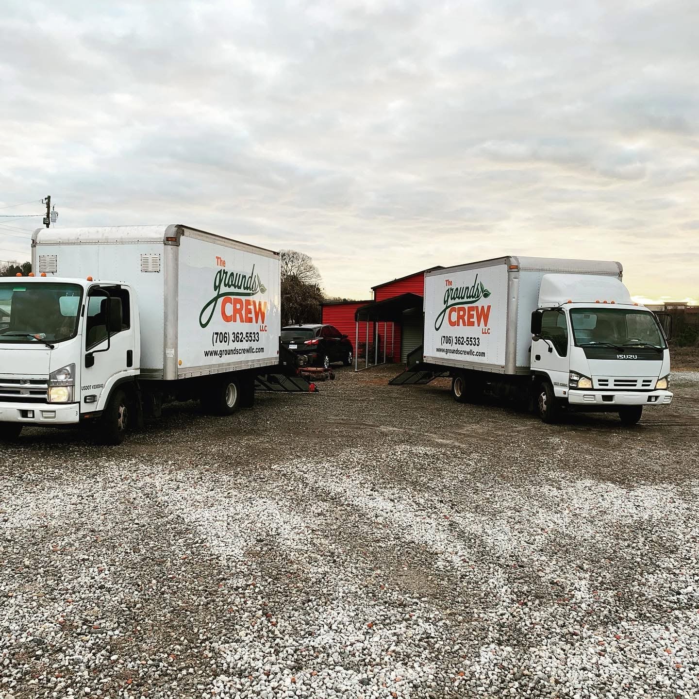 Two white moving trucks with 'The Grounds Crew LLC' logo parked on gravel outside a red building with a car trailer and a black car.