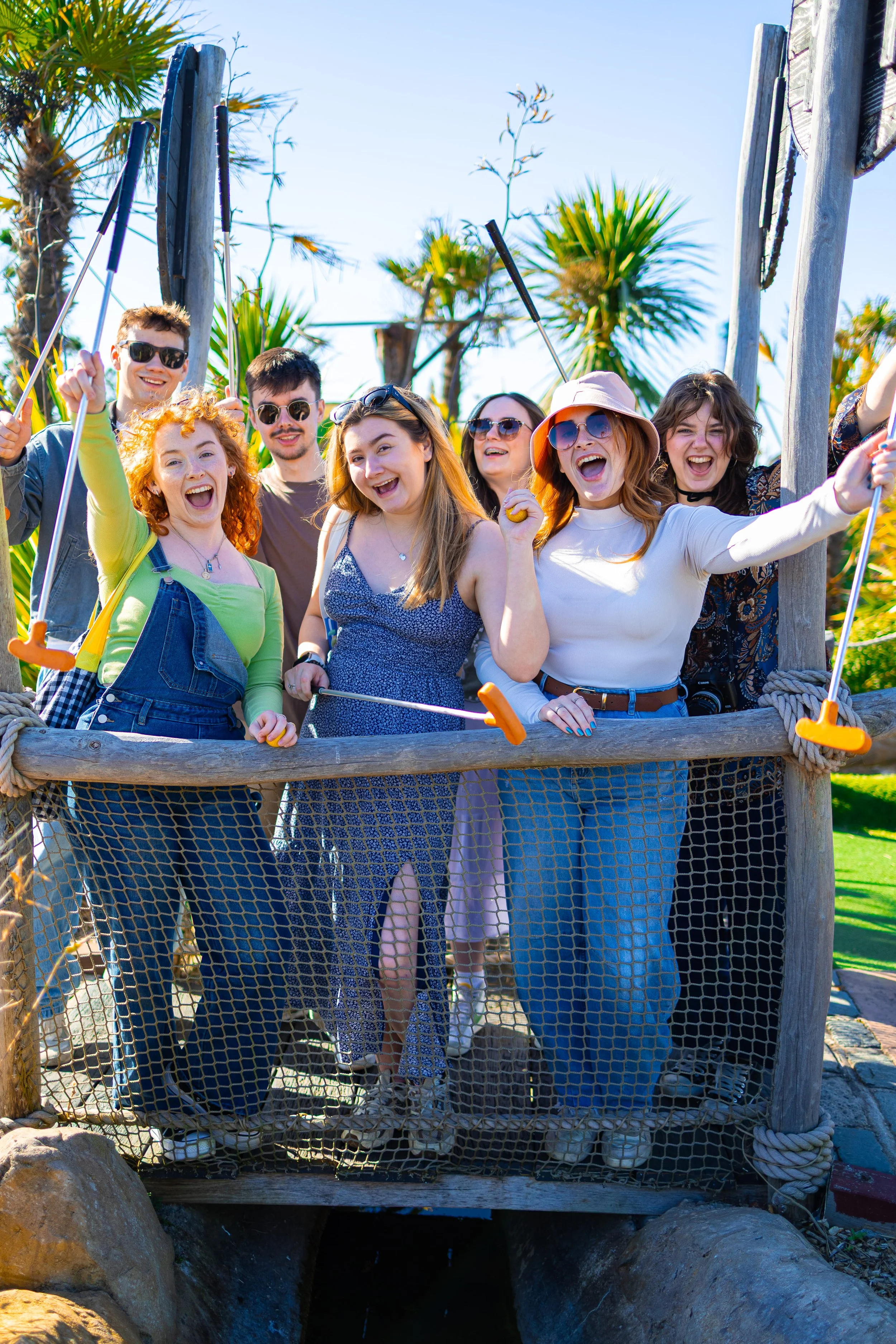 Group of people standing on a wooden bridge outdoors, smiling and posing for the photo, with palm trees in the background, wearing casual summer clothes and sunglasses.