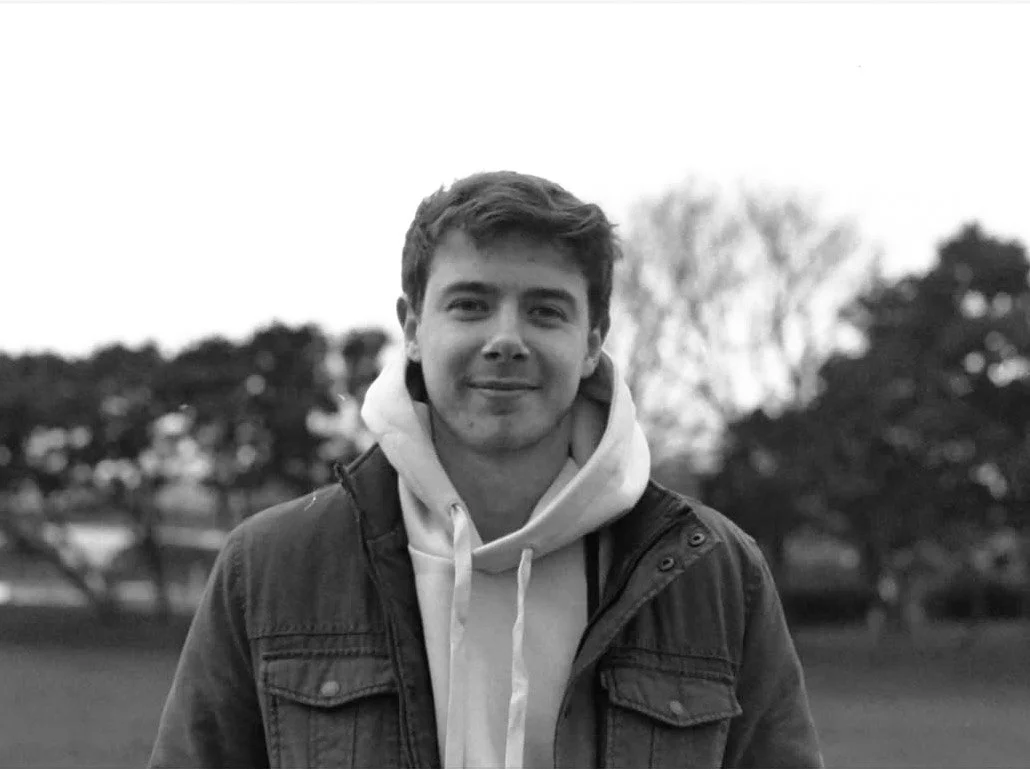 A young man with short, wavy hair, smiling, wearing a white hoodie and a dark jacket, standing outdoors with trees in the background on a cloudy day.