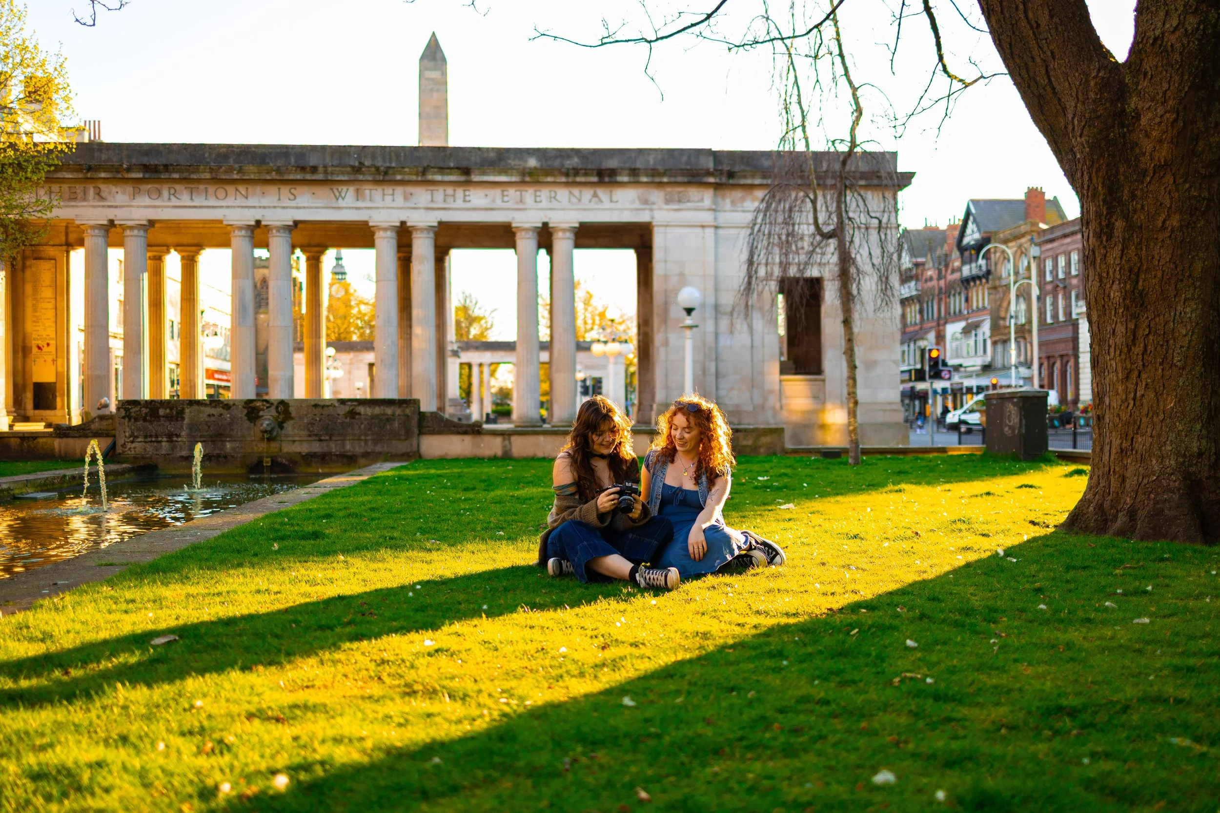 Two young women sitting on grass, looking at a camera, in a park with a historic building in the background, during sunset.