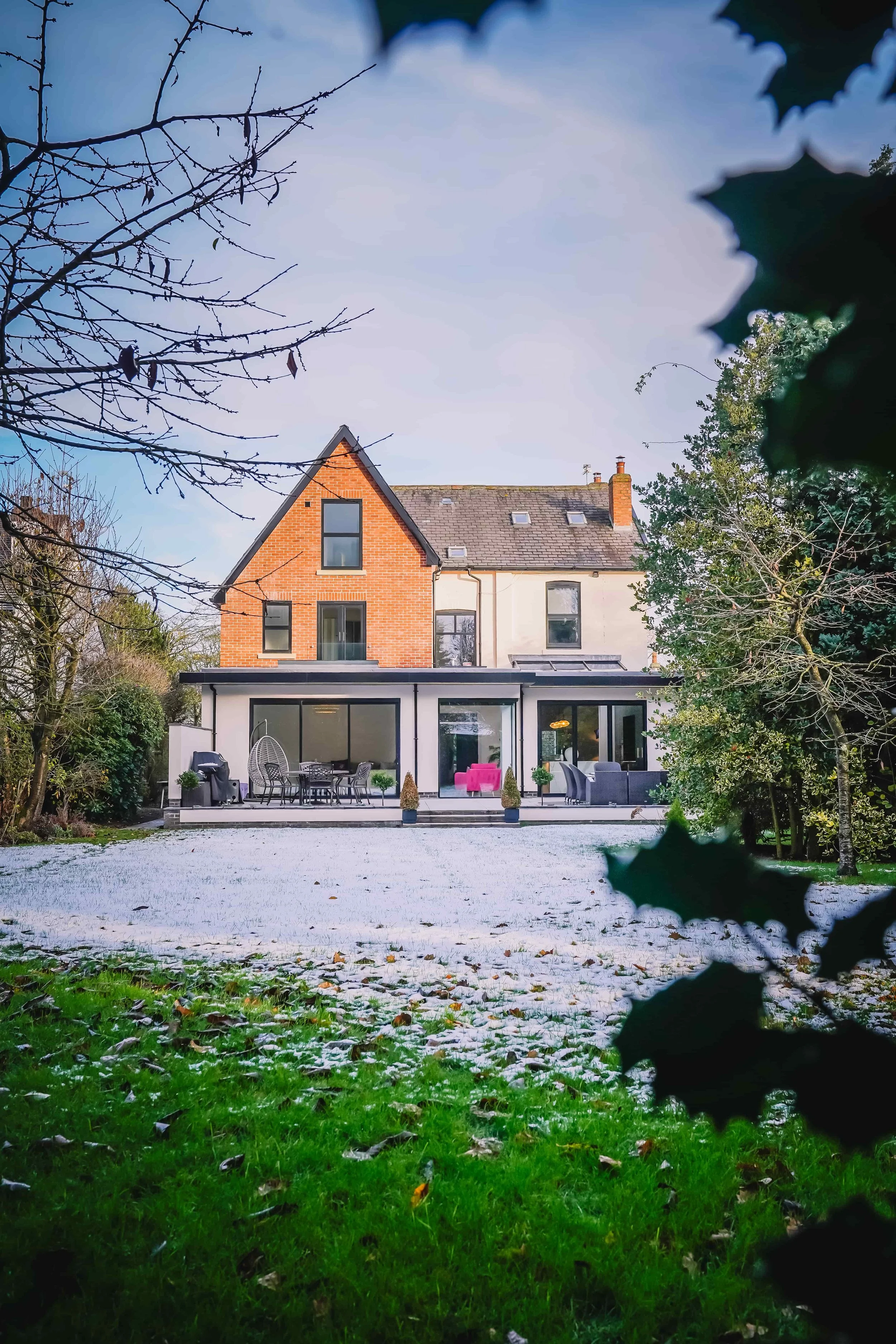 A house with a modern white extension at the back, surrounded by trees with some snow on the ground and a cloudy sky overhead.
