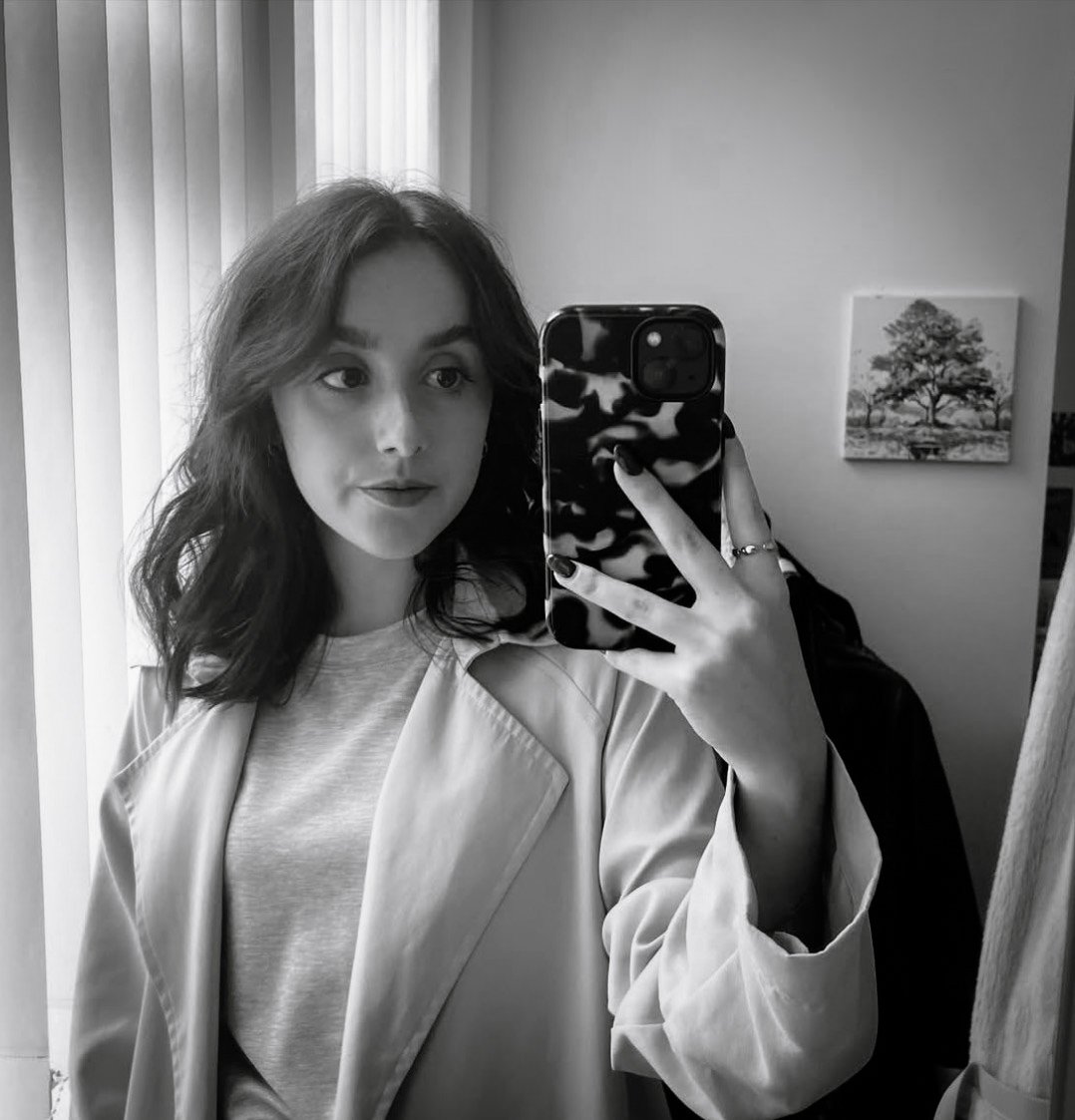 A young woman taking a mirror selfie with her phone in black and white. She has shoulder-length wavy hair, wears a light-colored jacket over a t-shirt, and has rings on her fingers. The background shows vertical blinds, a wall with a framed tree painting, and a clothing item hanging on a hook.