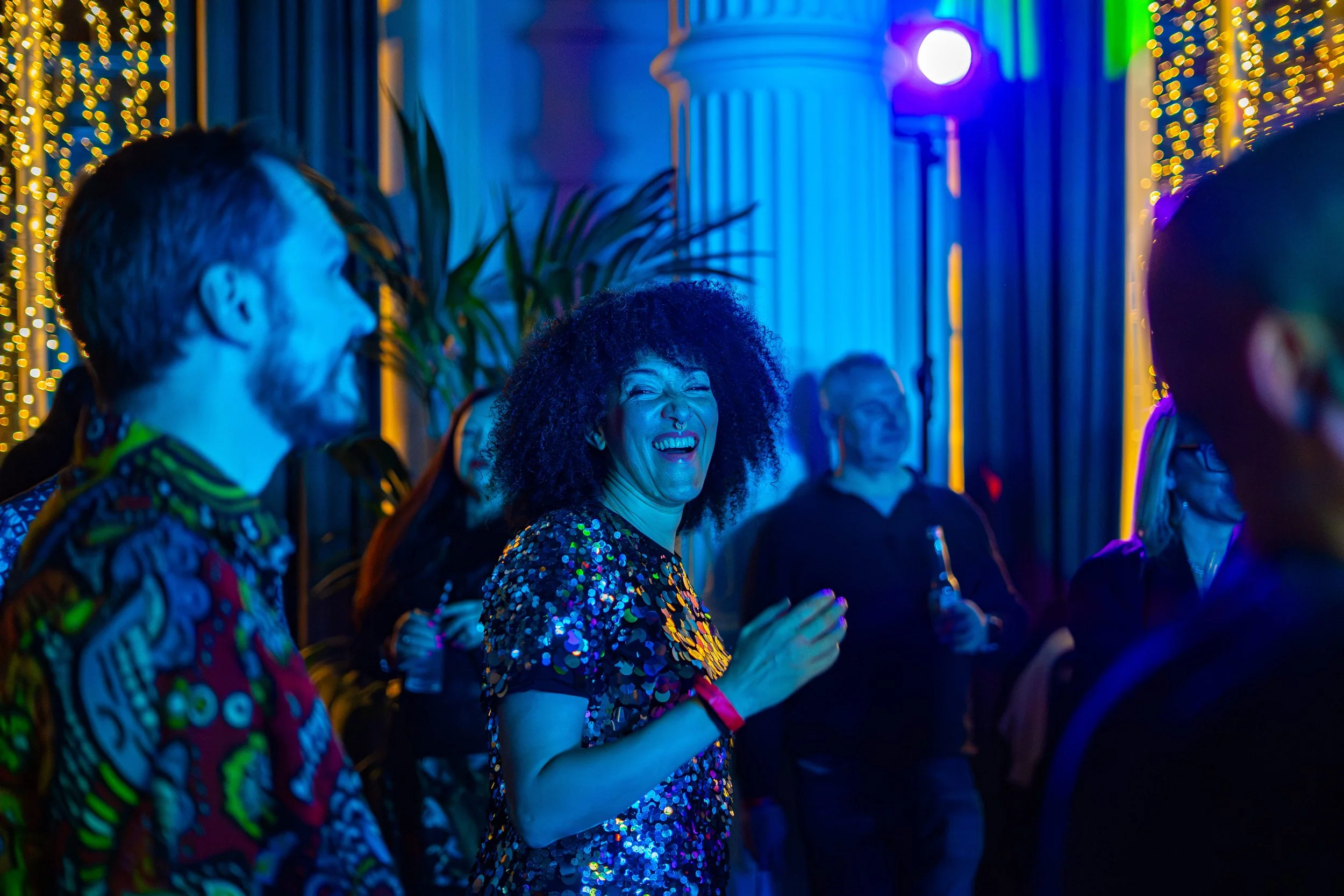 People dancing and laughing at a party under colorful, festive lighting decorated with string lights.
