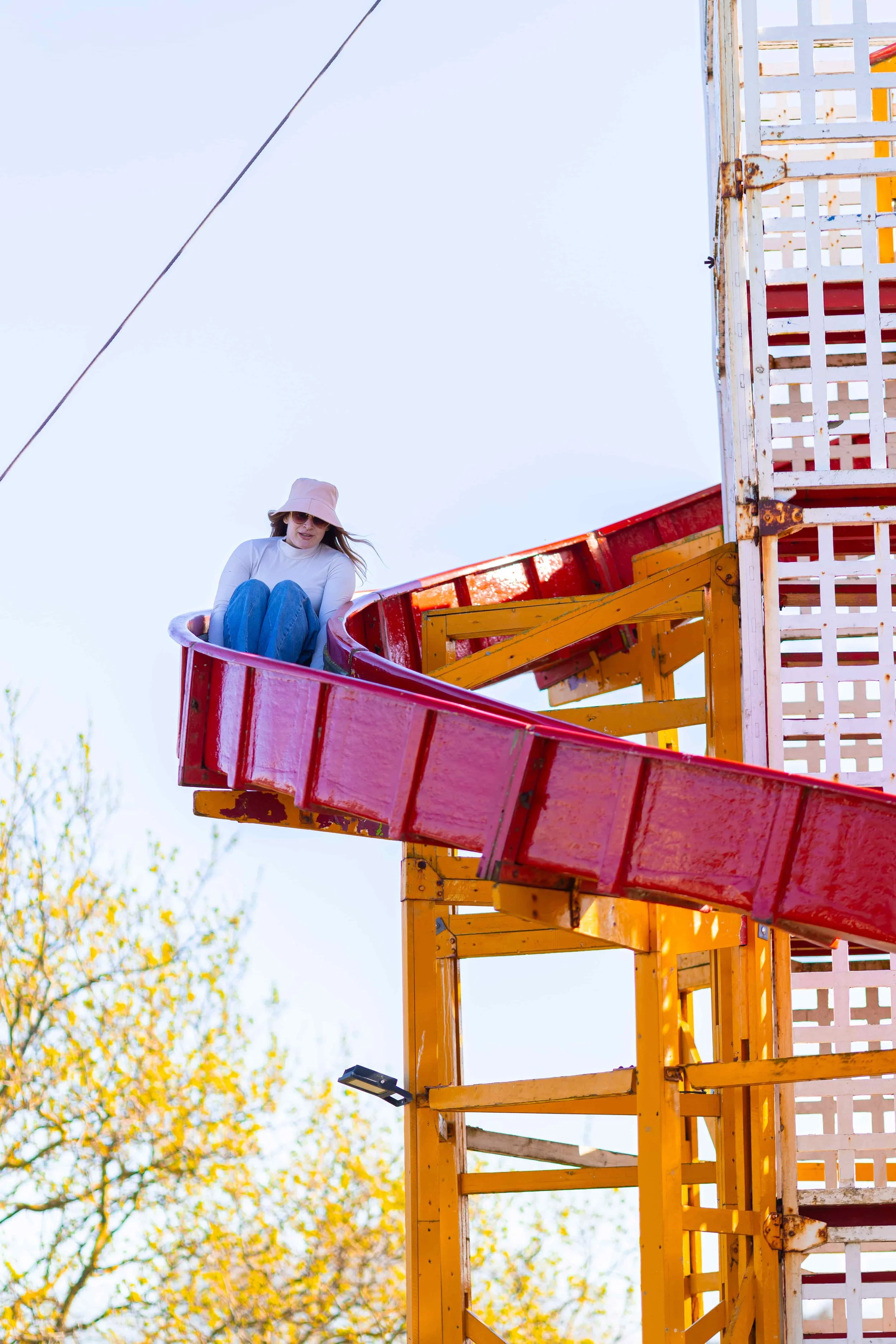 A woman wearing sunglasses, a light pink hat, a white long sleeve top, and jeans, sitting on a red slide at a playground, with a yellow and brown play structure and the sky in the background.