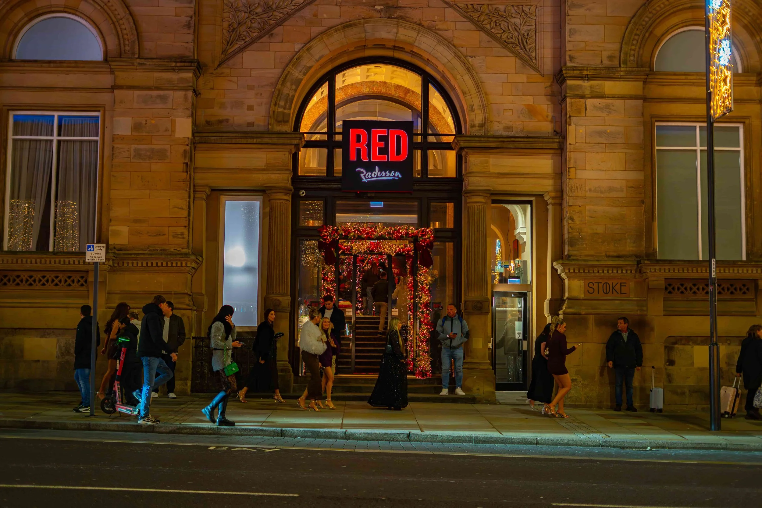 Nighttime scene outside a historic stone building decorated with Christmas lights. People walking and standing in front of the entrance, which is illuminated and decorated with red ribbons and greenery. A large sign reads 'RED Radisson' above the entrance.