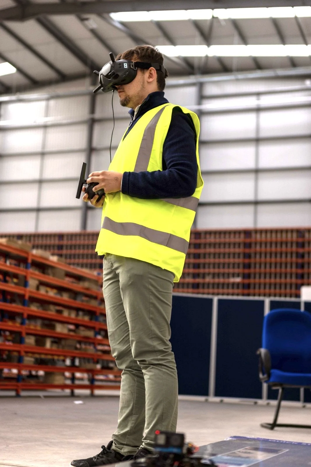 A man wearing a yellow safety vest and virtual reality goggles operating a remote control in an industrial or warehouse setting.