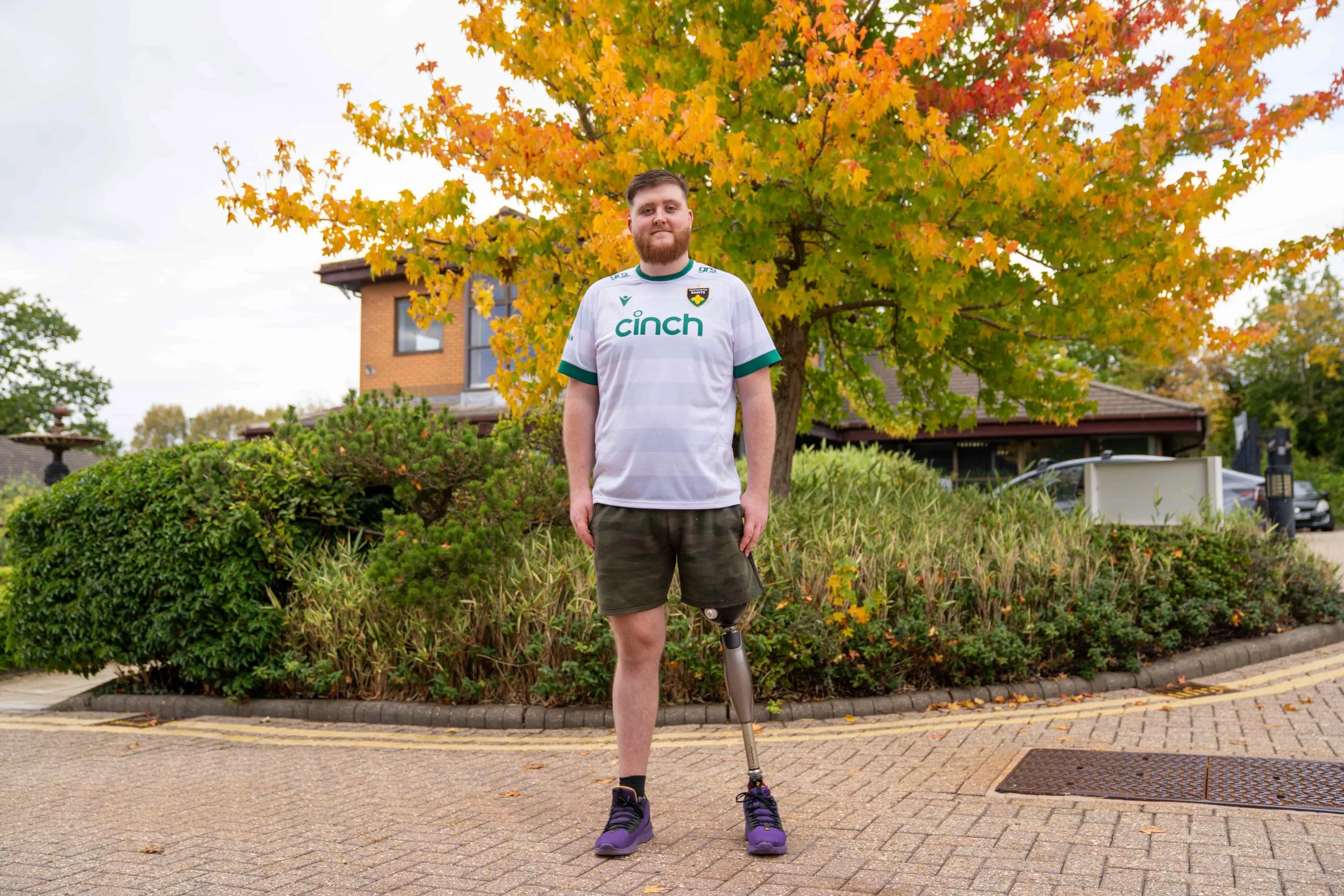 A young man with a beard, wearing a white sports jersey and shorts, posing with a prosthetic limb on his right leg underneath a large tree with yellow and orange autumn leaves, in a suburban neighborhood.