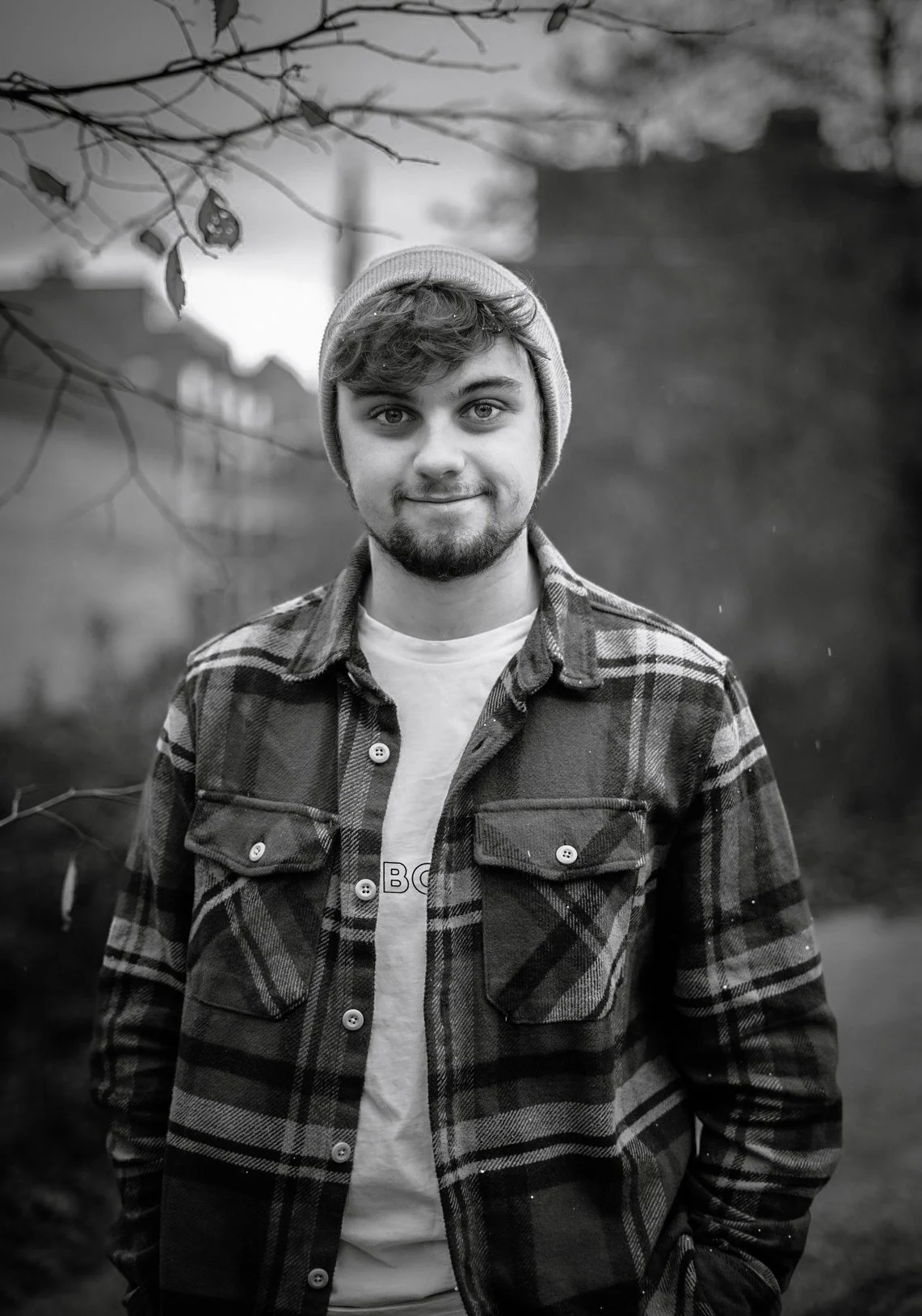 A young man with a beard and short hair wearing a beanie, a plaid shirt, and a t-shirt stands outdoors on a cloudy day. Leaves and branches are visible in the foreground and background.