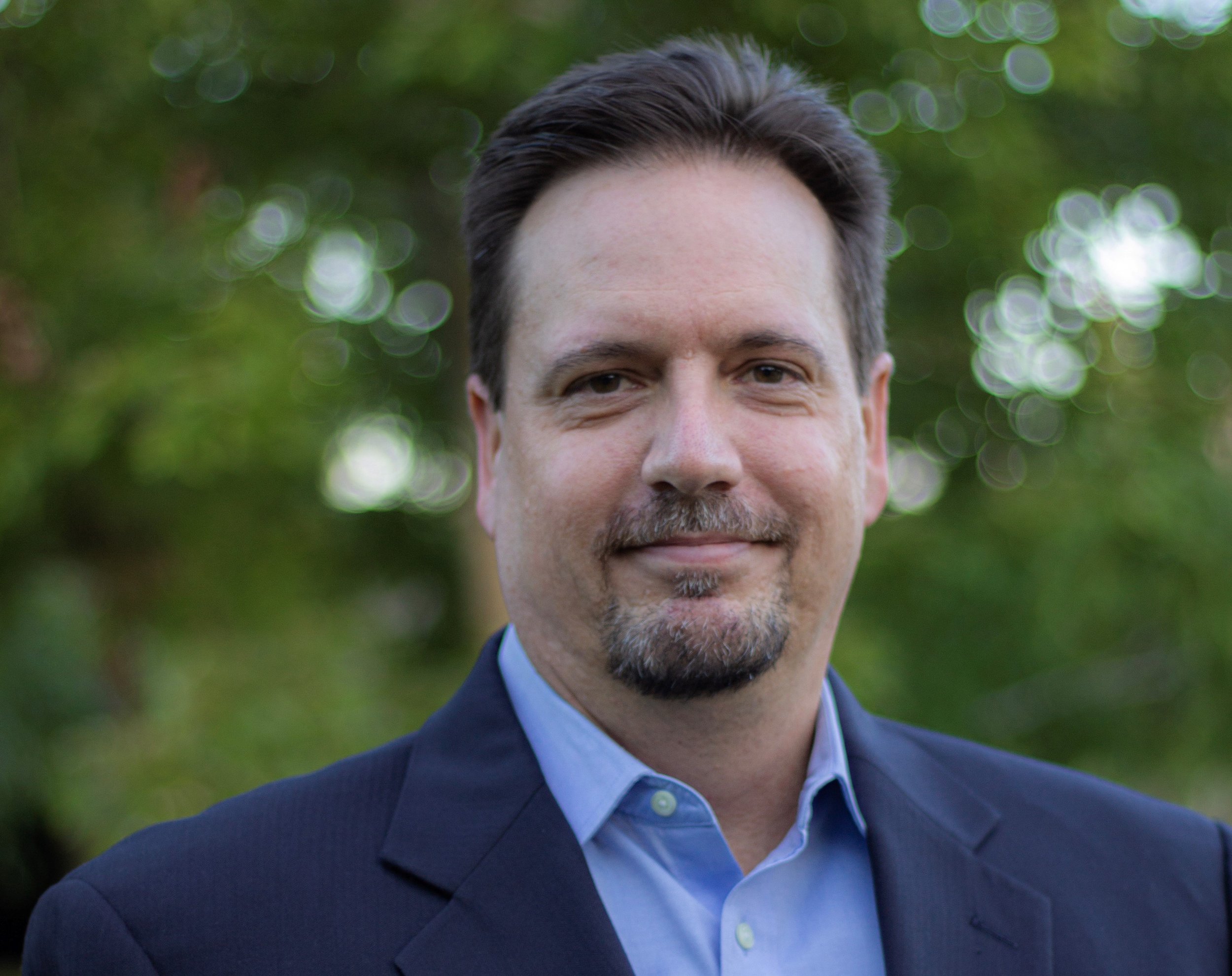 A man with dark hair, a mustache, and a beard, wearing a dark suit jacket and a light blue collared shirt, stands outdoors with a blurred green background and bokeh effect.