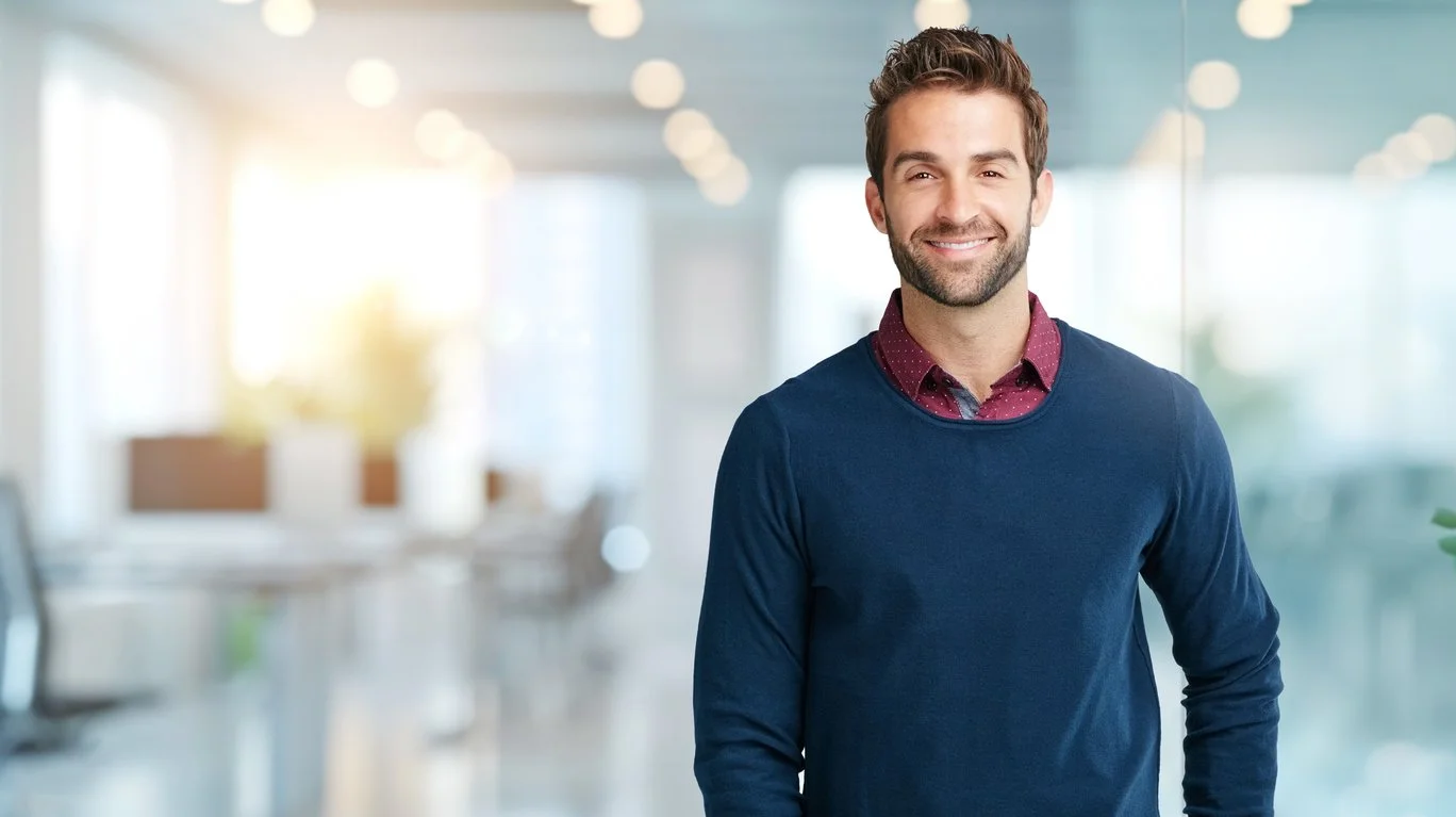 A smiling man with brown hair and a beard, wearing a blue sweater over a red collared shirt, standing in a bright, modern office.