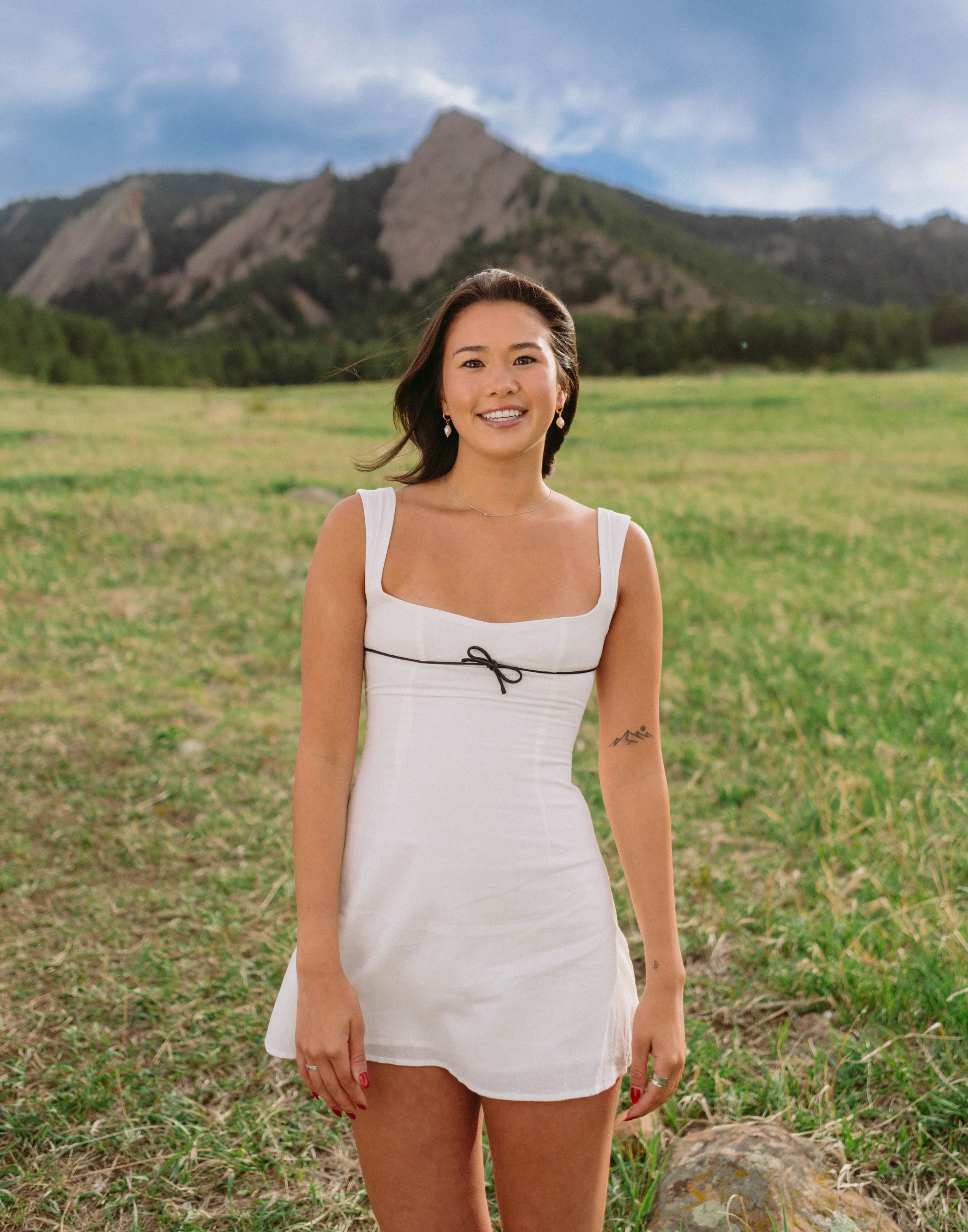 “CU Boulder senior portrait in white dress standing in green field with Flatirons”