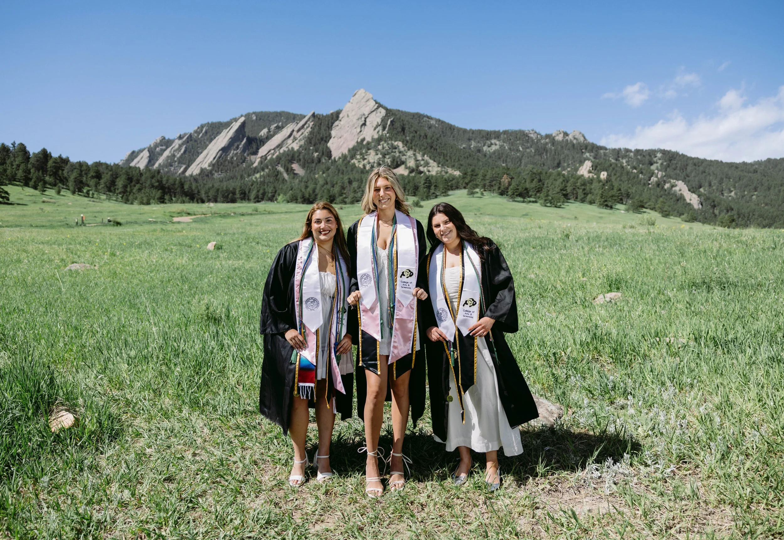 Three CU Boulder grads in cap and gown posing in a bright green Chautauqua meadow with the Flatirons behind them, classic Boulder graduation scenery.