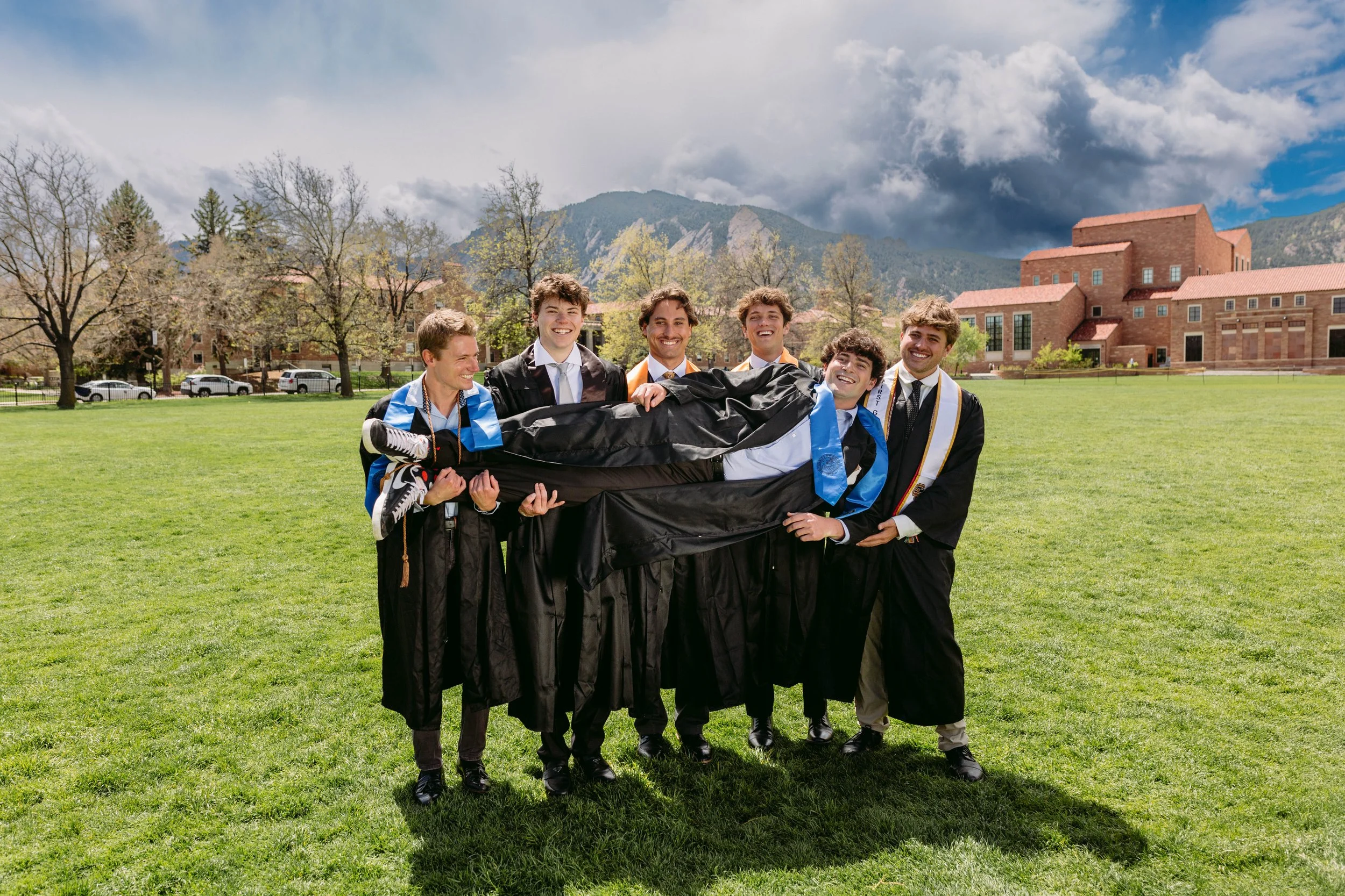 Fun CU Boulder group graduation photo of six friends at Farrand Field with the Flatirons behind them, showing candid and energetic moments.