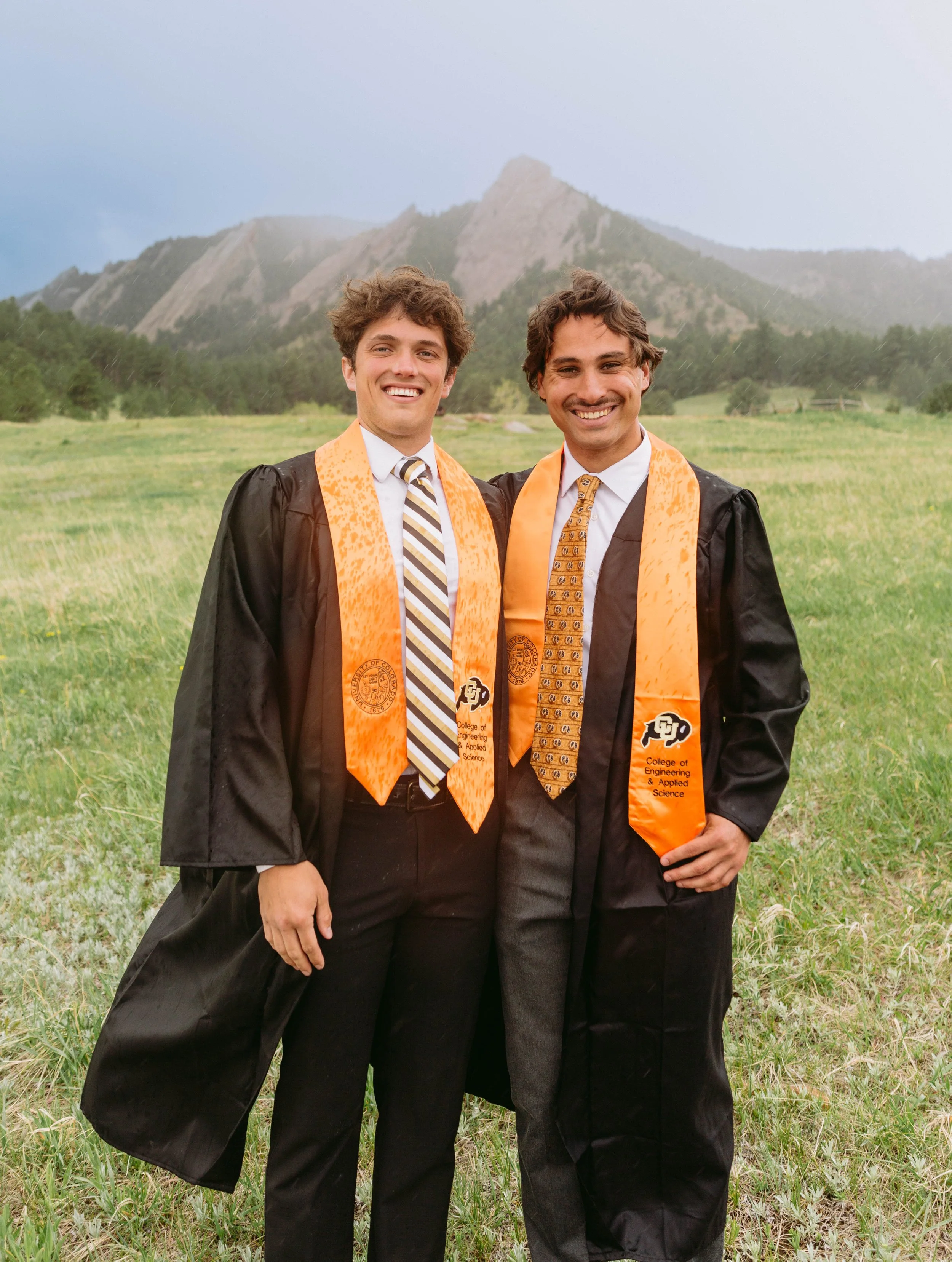 Two men in graduation caps and gowns standing in a grassy field with mountains in the background, smiling at the camera.