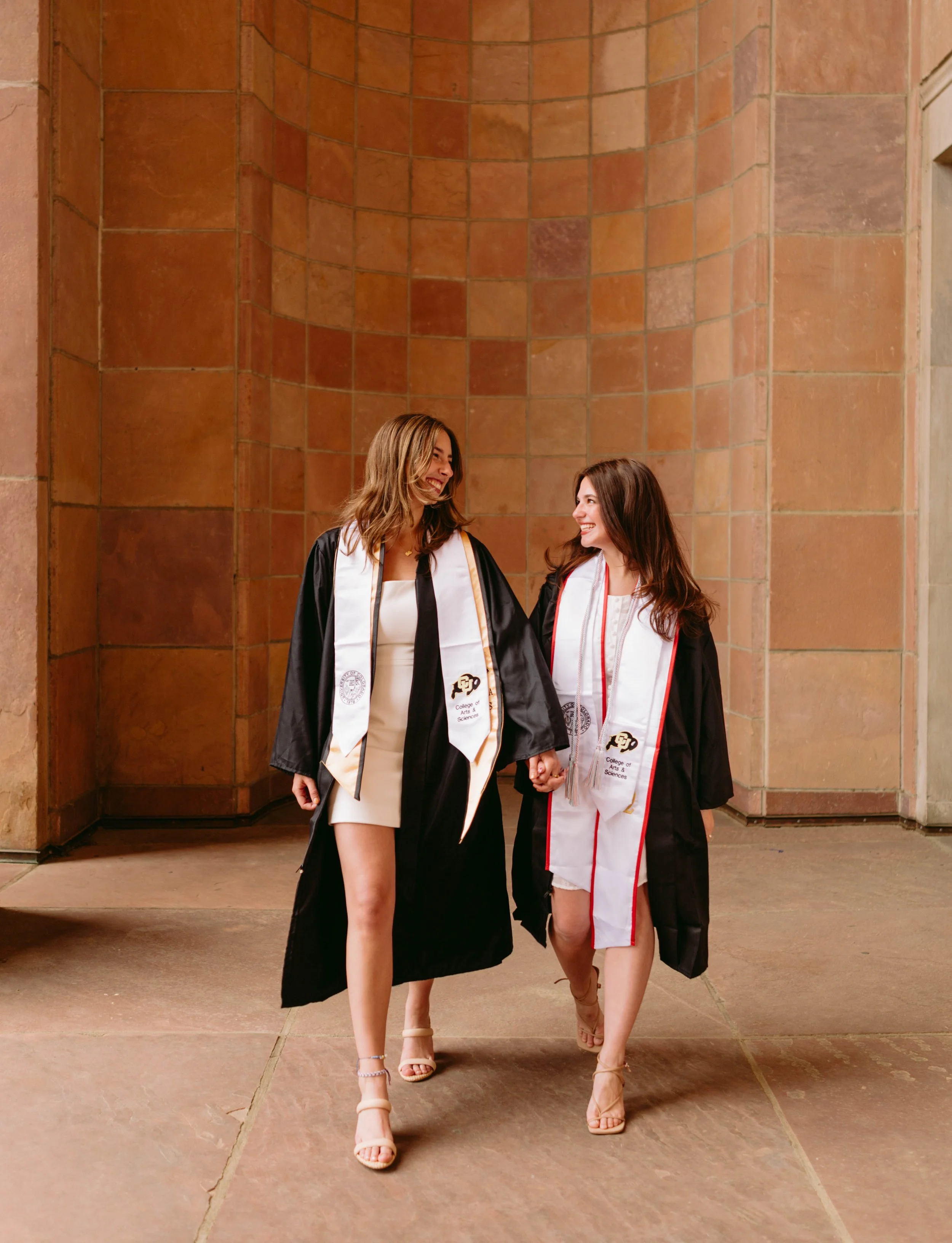 Two CU Boulder seniors walking together under campus archway in graduation attire