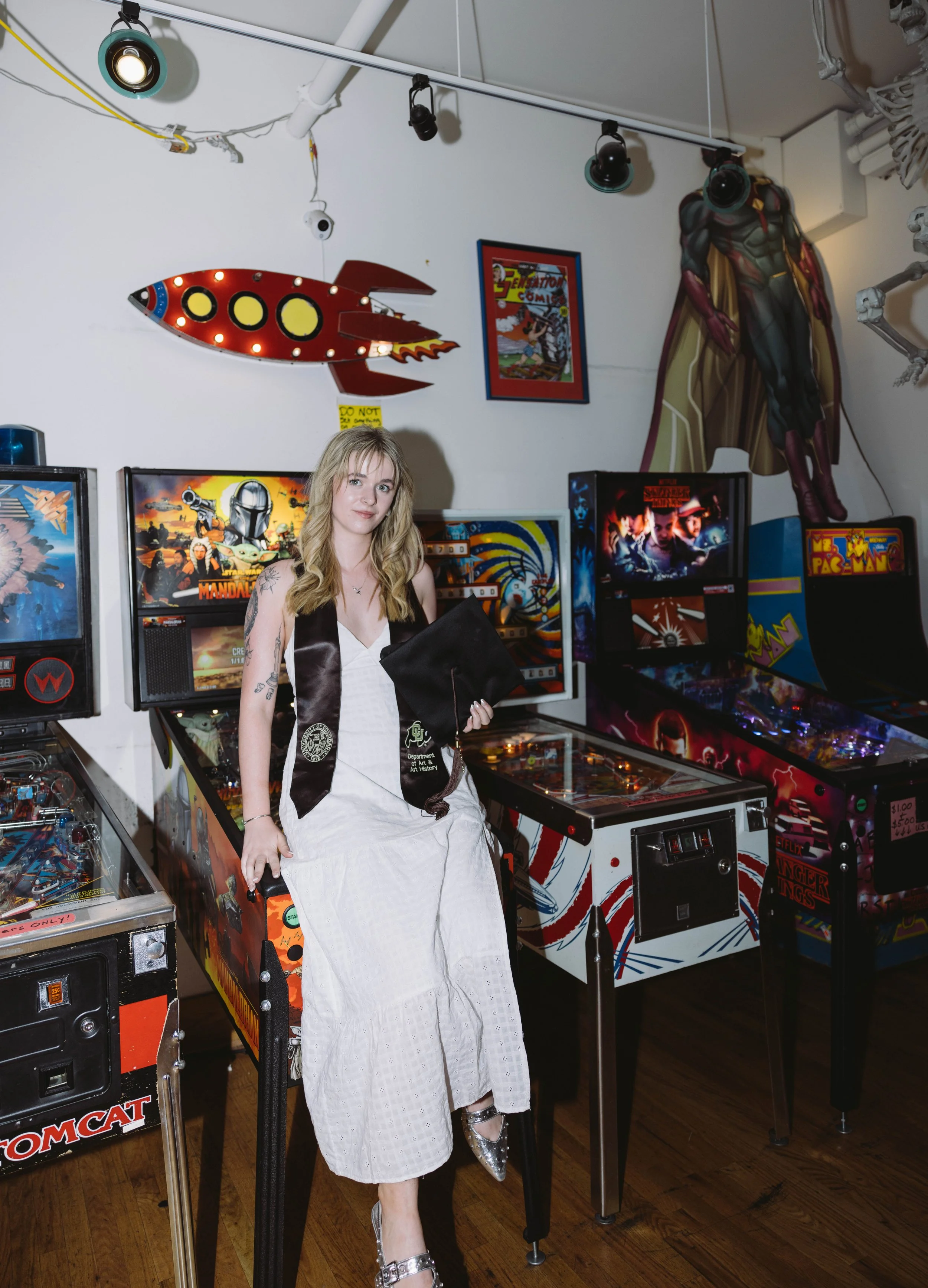 Female CU Boulder grad at a pinball arcade in a white dress with her stole