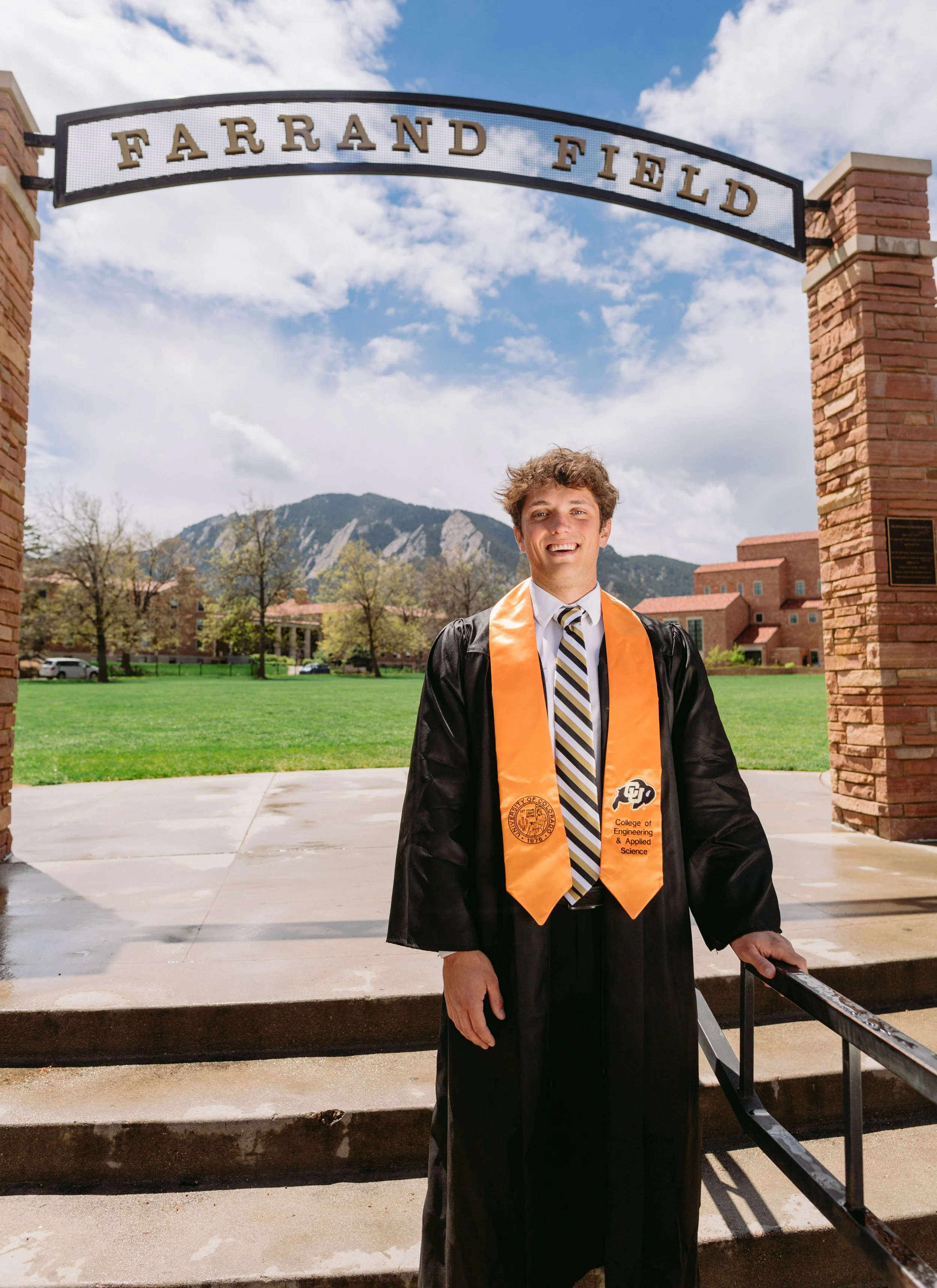 CU Boulder senior portrait in cap and gown at Farrand Field with Flatirons in background