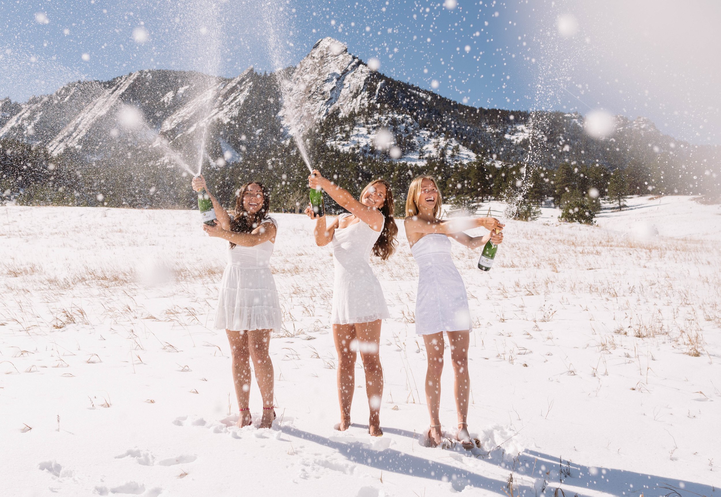 Three girls in white dresses in front of the flatiorns in Boulder colorado, popping champagne to celebrate graduating from the university of colorado