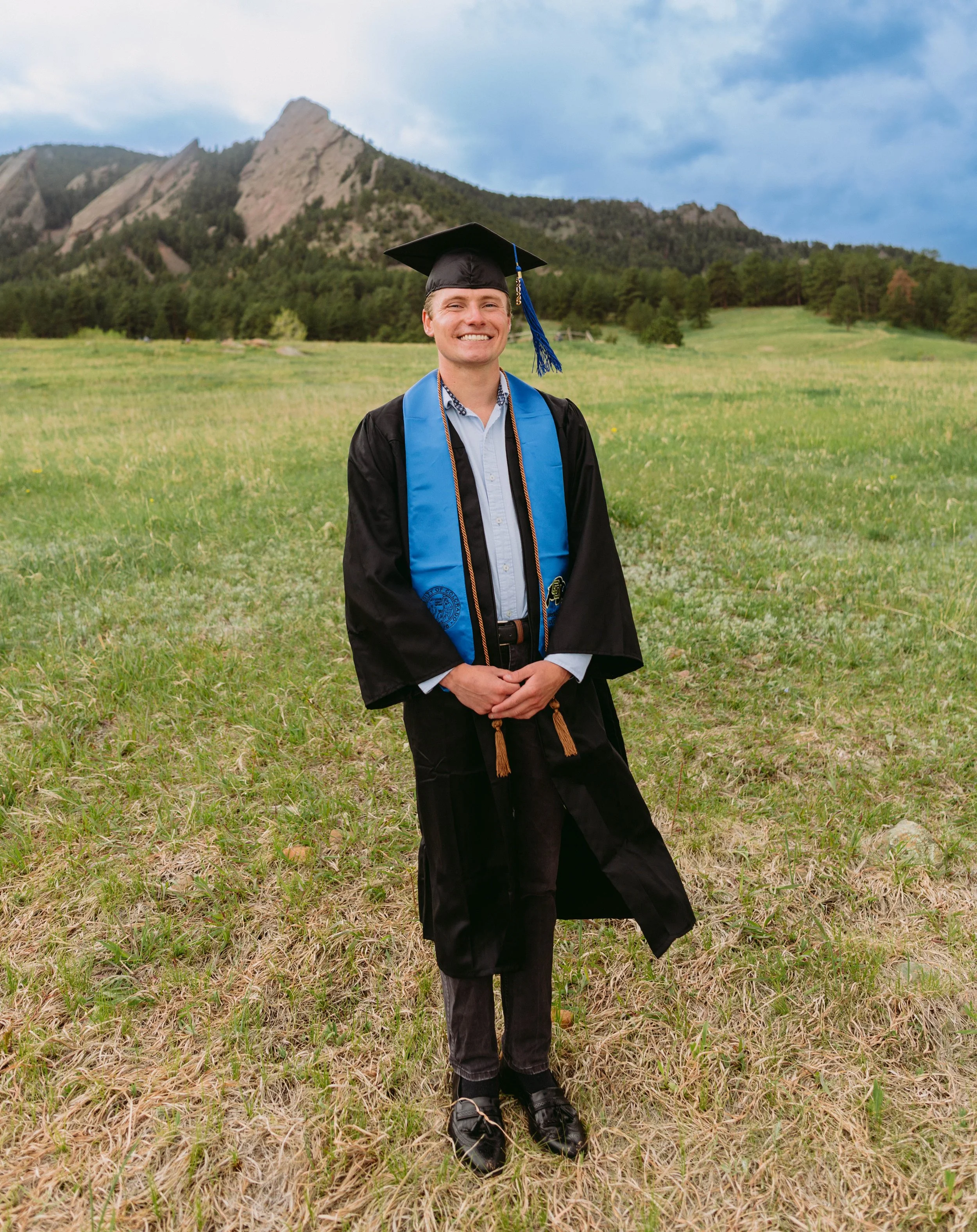 Senior graduation portrait of a CU Boulder graduate standing in a Flatirons meadow at Chautauqua Park, featuring classic cap-and-gown styling and clean natural light.