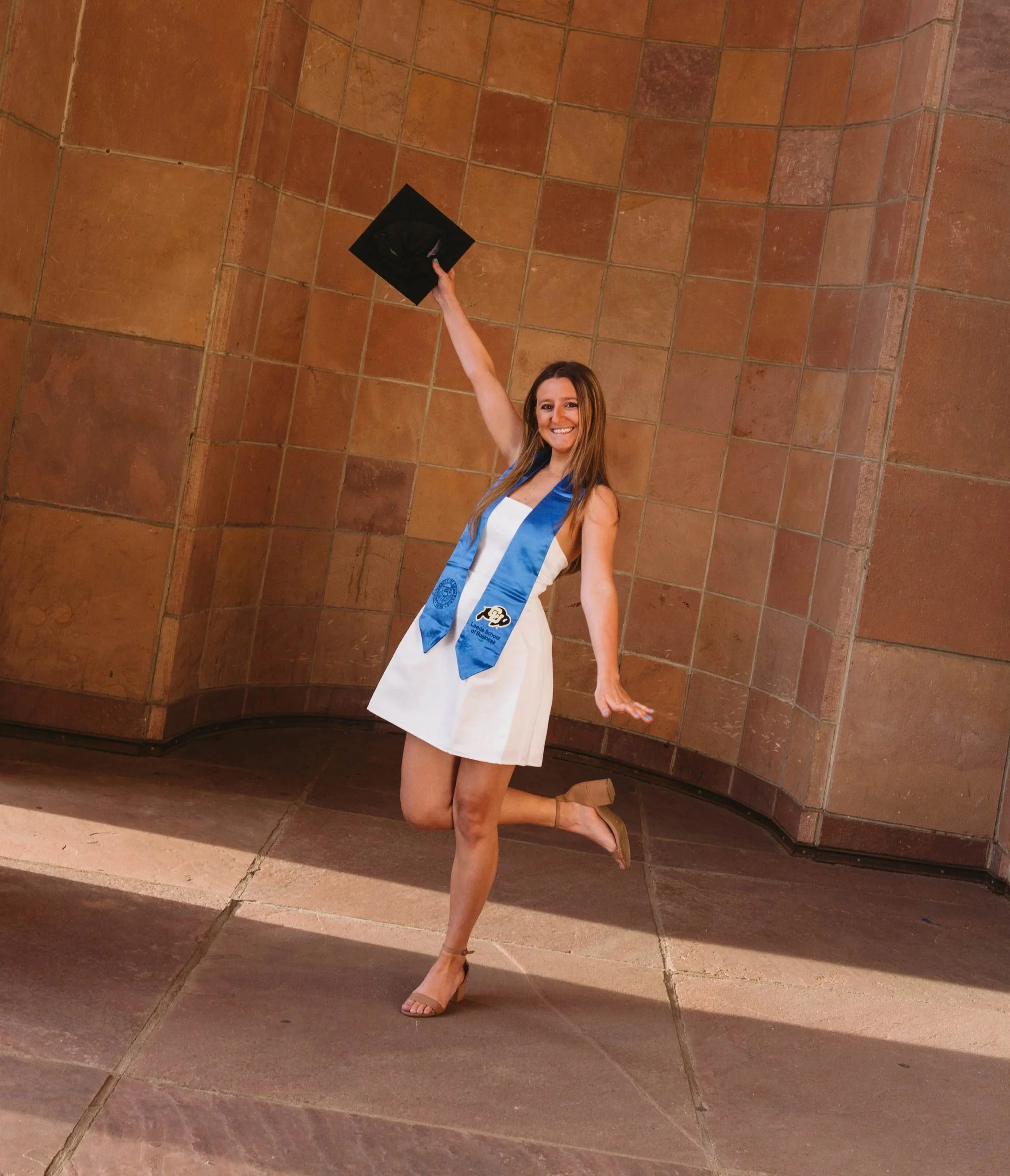 Woman in white dress, holding her cap, celebrating her graduation from the University of Colorado boulder.