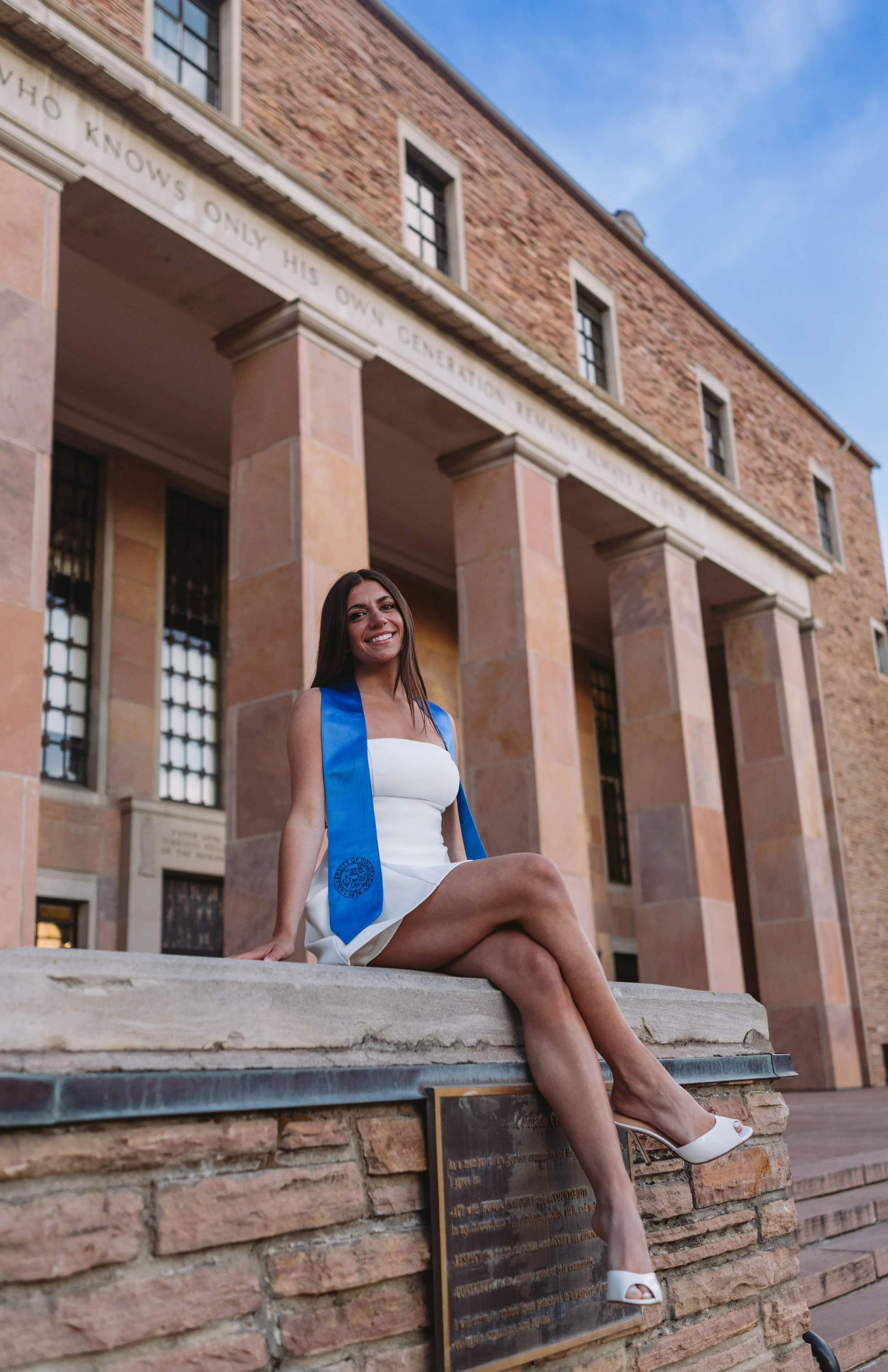 CU Boulder senior sitting on campus steps wearing graduation stole and white dress