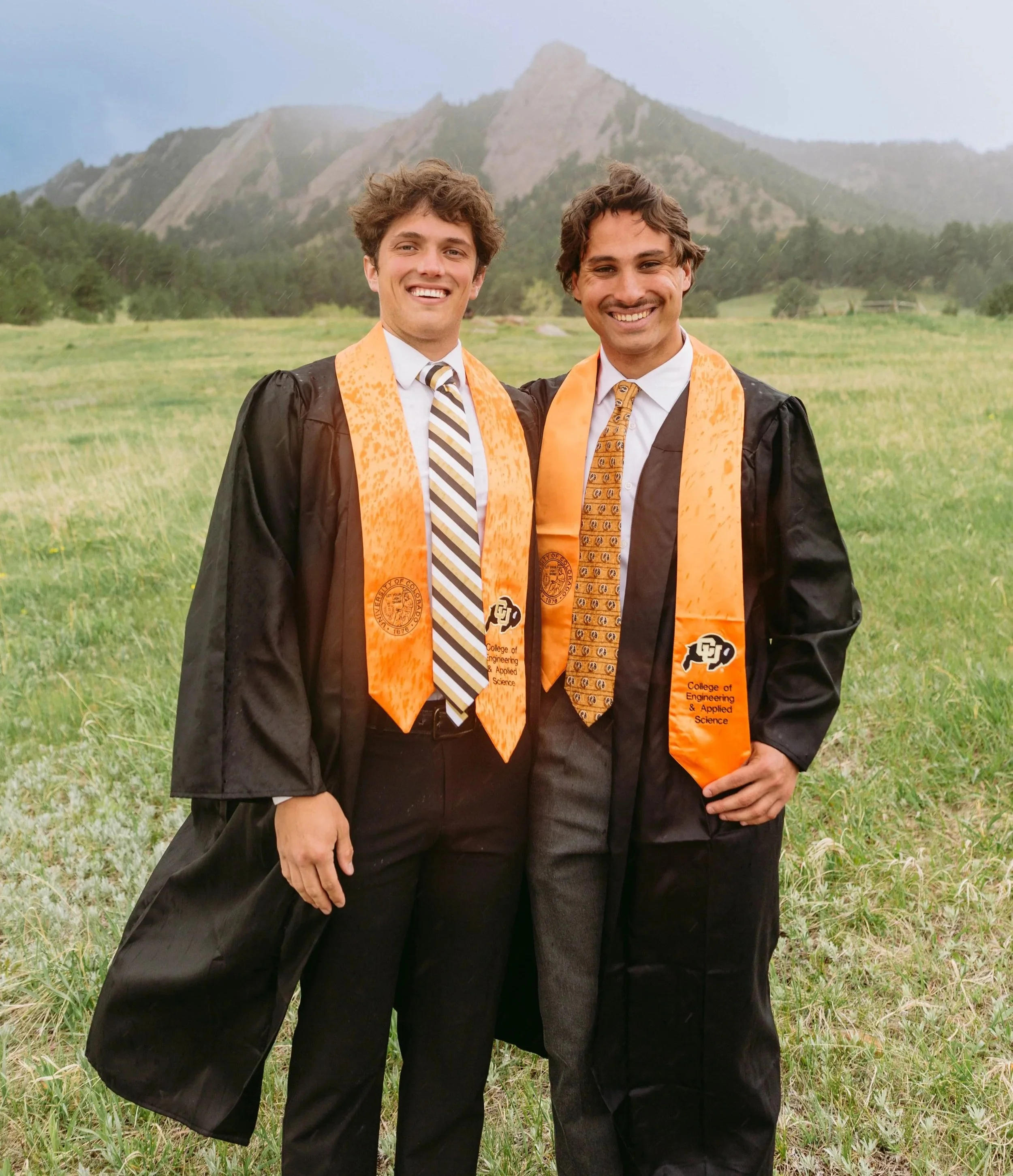 Two young men in graduation caps and gowns, standing outdoors on a grassy field with mountains in the background.