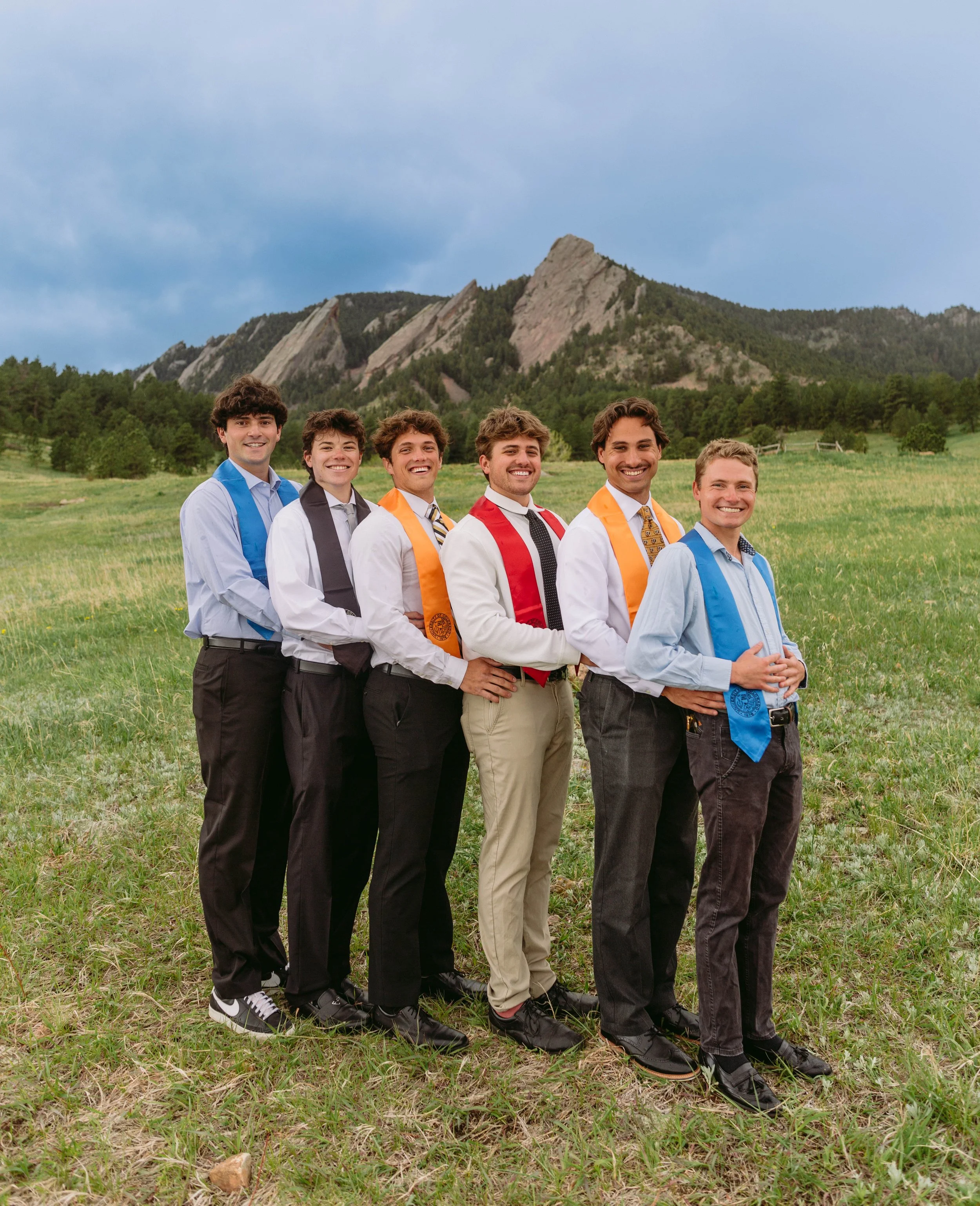 Group of CU Boulder male graduates posing together in caps and gowns with Flatirons