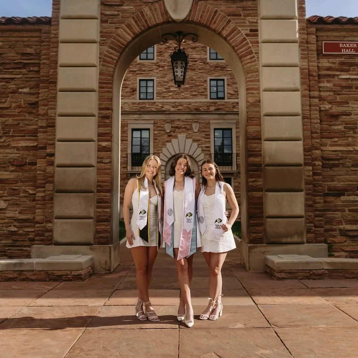Three women in white dresses and graduation stoles standing together on a brick walkway in front of a brick building with an arched entrance, celebrating graduation, smiling, sunny day.