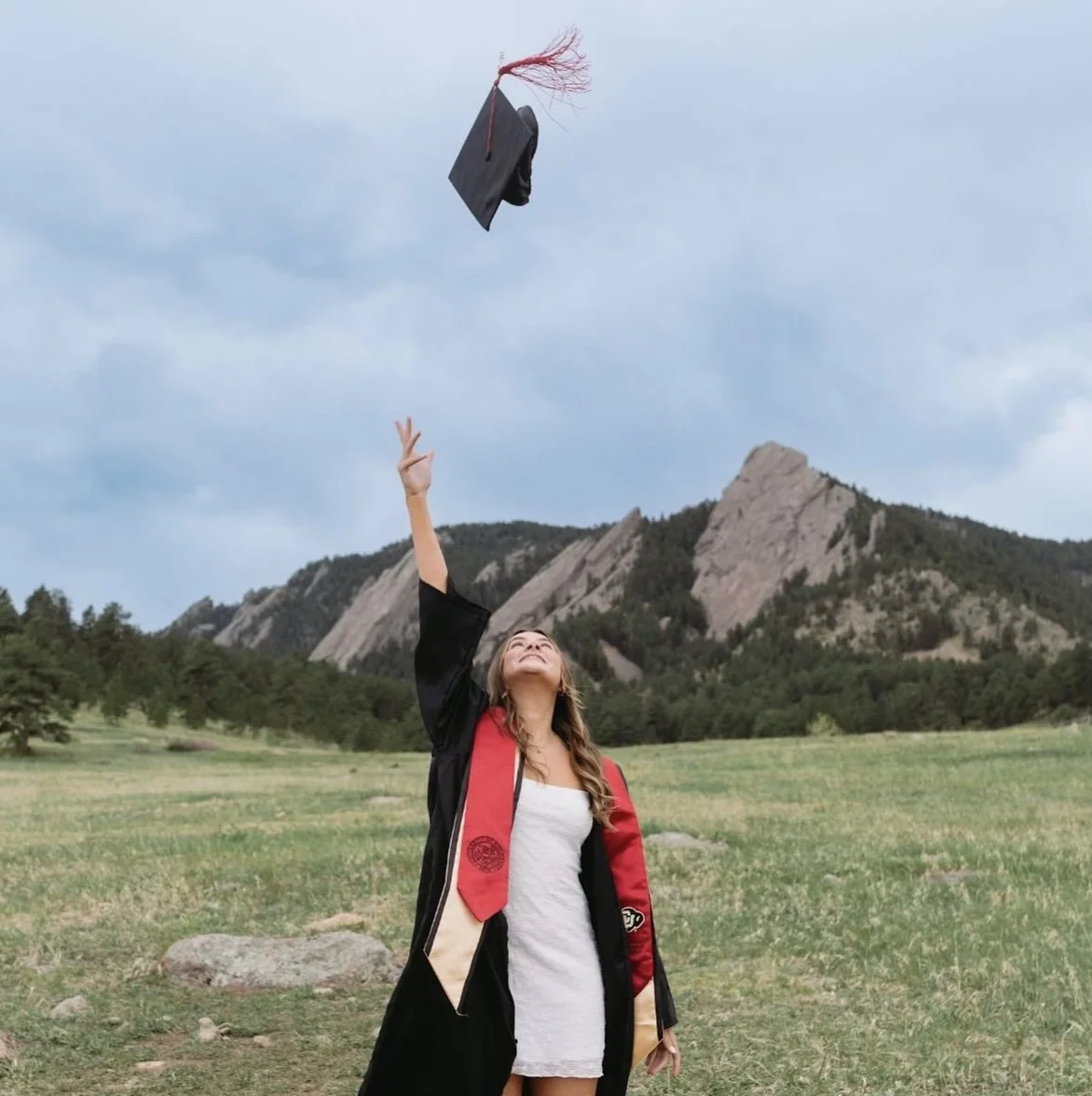 A woman in graduation attire throwing her cap in the air outdoors with mountains and trees in the background.