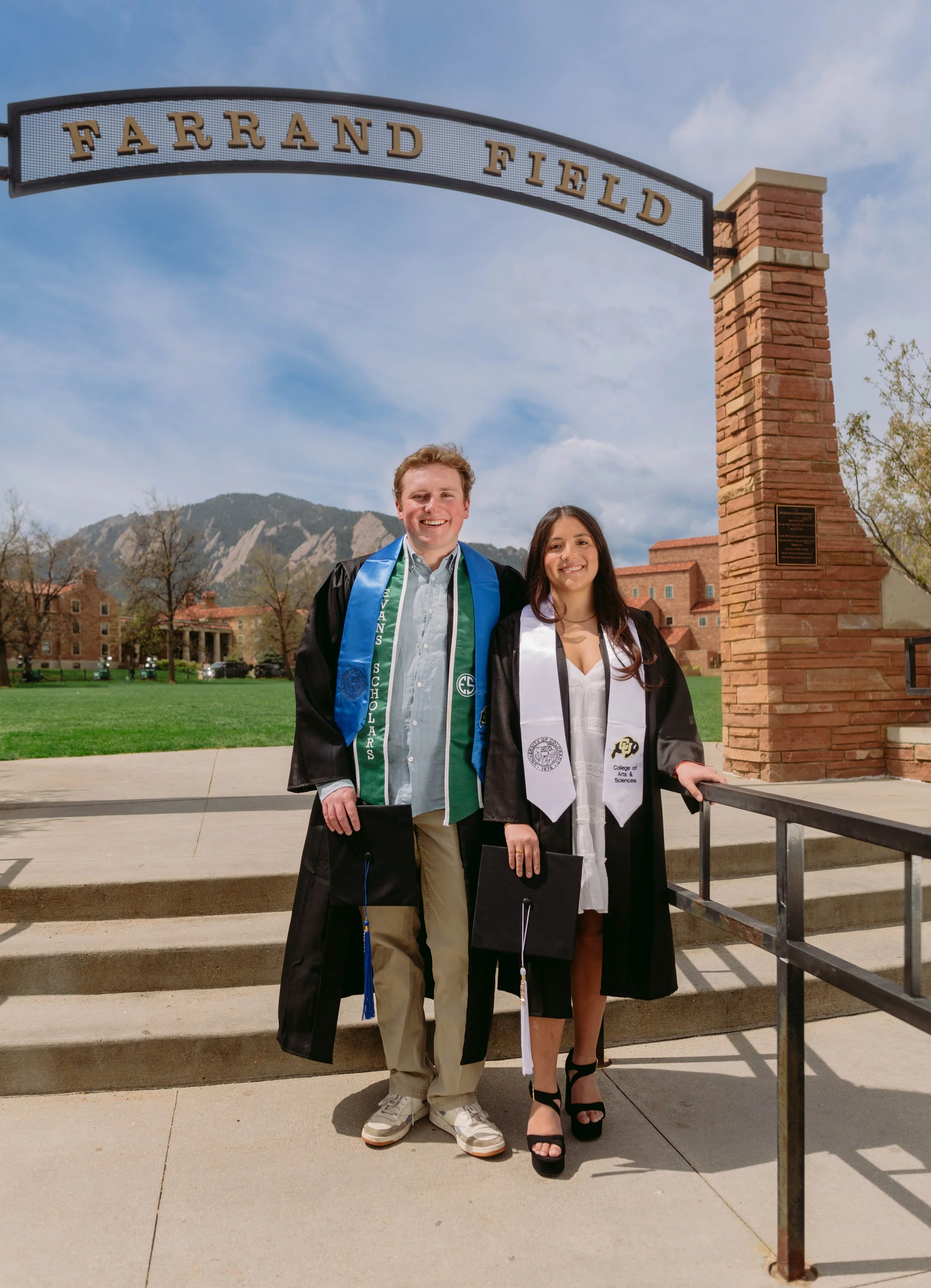 Two graduates wearing caps and gowns standing under a sign that reads 'Farrand Field' at a university campus, with a mountain in the background.