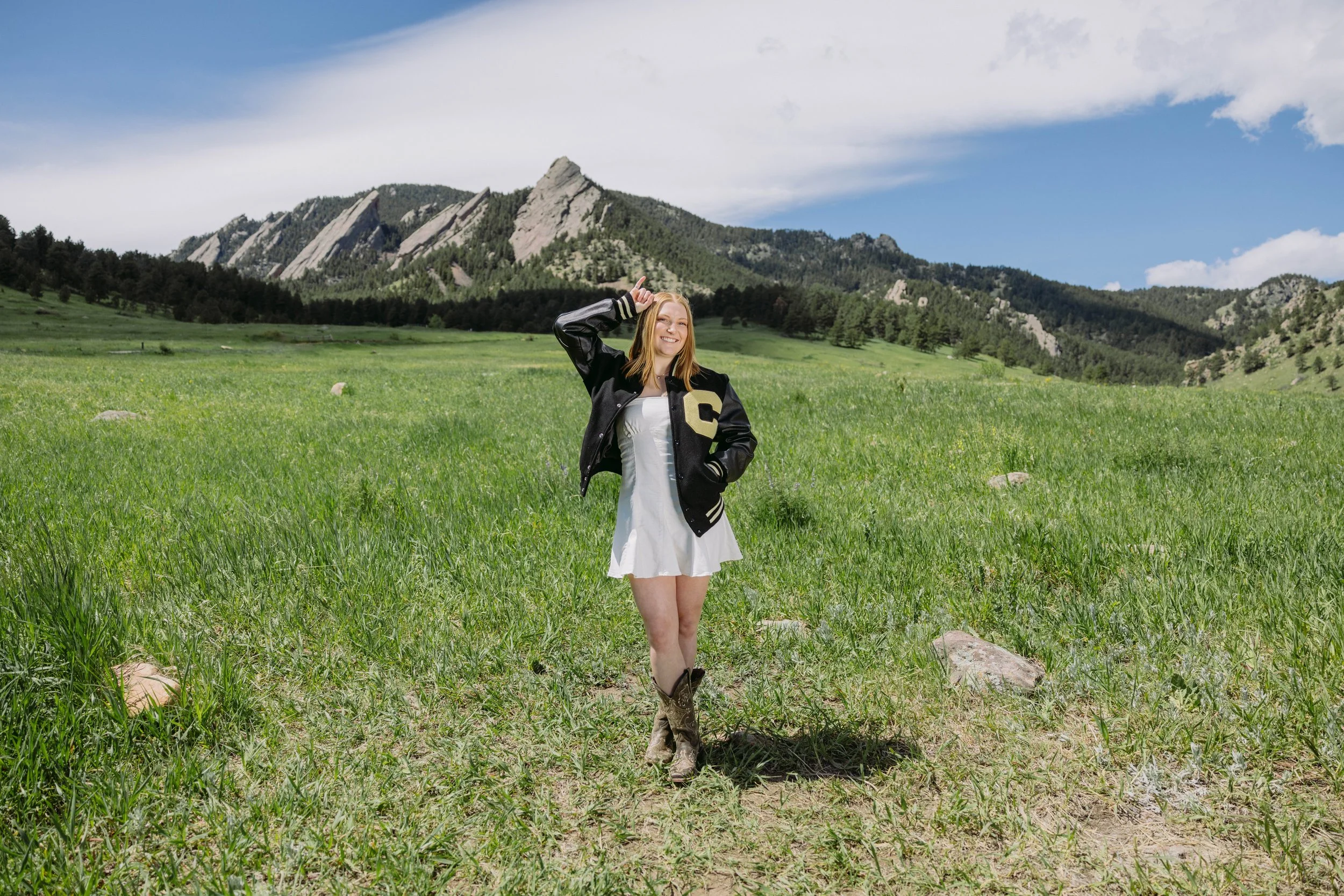 CU Boulder senior portrait in green field wearing letterman jacket with Flatirons