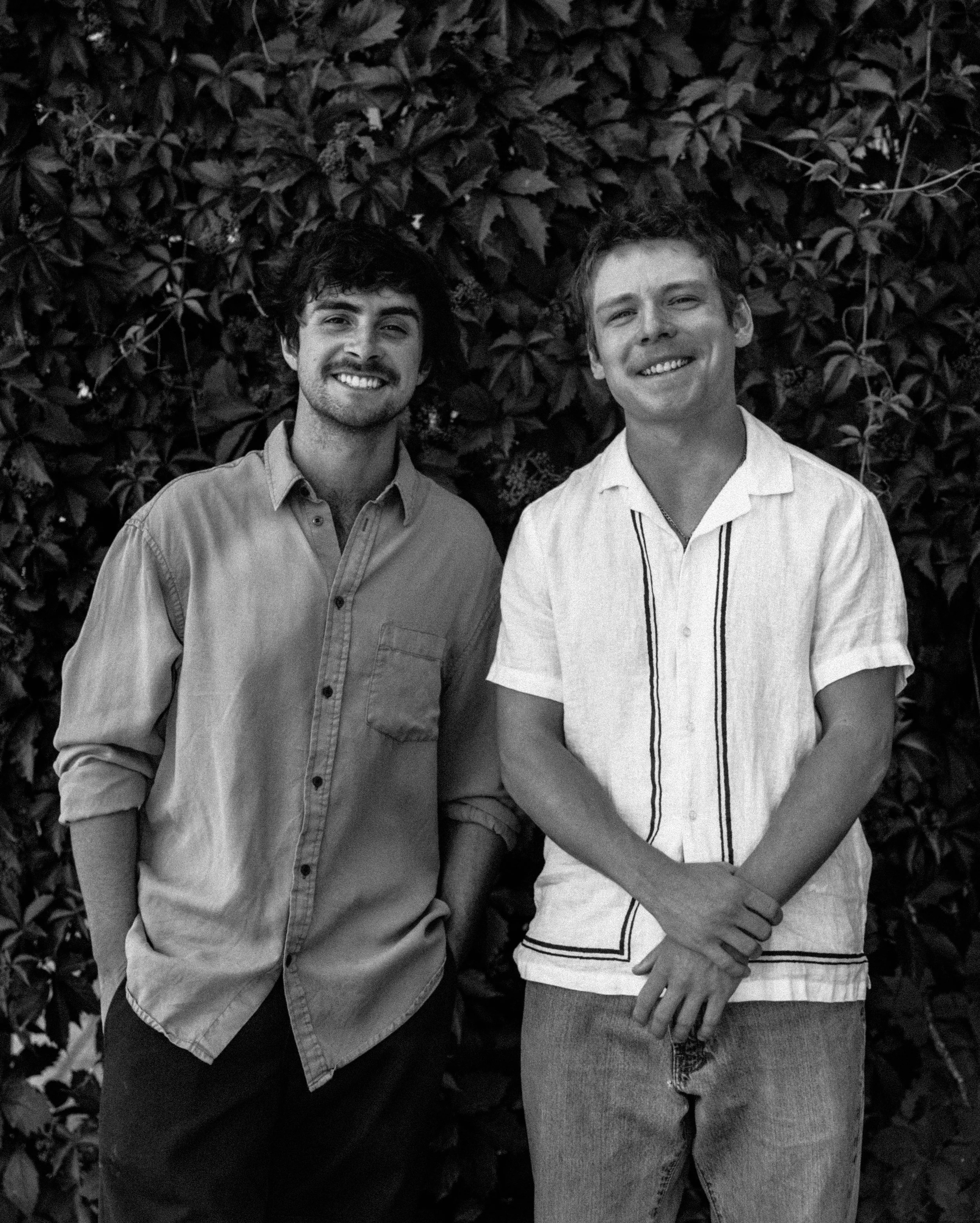 Two young men standing side by side in front of a leafy shrub, smiling at the camera.